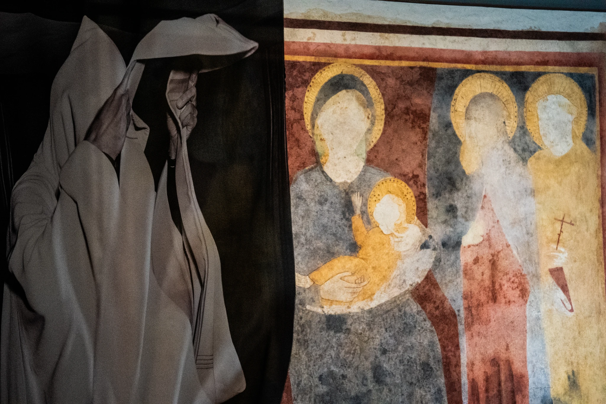 Immagine che mostra affiancati un pannello con una figura avvolta in un drappo chiaro, raffigurata mentre solleva il tessuto sopra il capo, e un affresco antico con tre figure aureolate. Nell’affresco, una figura centrale tiene in braccio un bambino con aureola, mentre le altre due figure sono in piedi a lato. I colori dell’affresco sono attenuati dal tempo, con toni di rosso, blu e ocra.