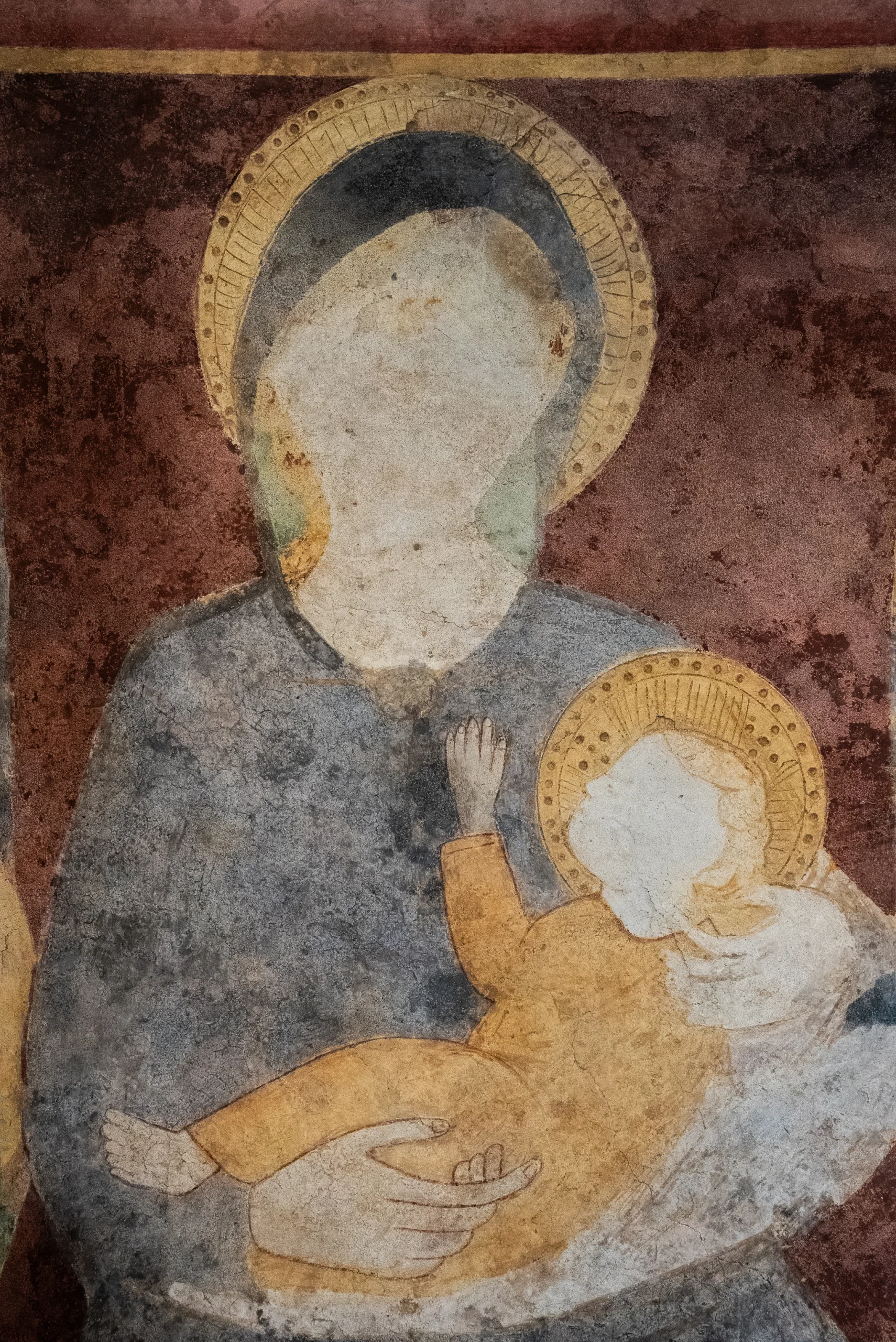 In un'immagine fotografica, che ritrae l'interno di un'aula, si scorge un affresco che raffigura una Madonna con Bambino su sfondo marrone sfumato sul giallo ocra e grigio. La Madonna, che indossa un mantello di colore grigio, si trova in primo piano e tiene il Bambino, di colore giallo miele, tra le braccia. Sulla sinistra si intravedono scheletri di animali di colore bianco. Sulla destra si distinguono alcuni elementi di colore giallo miele, tra cui un telo e una sedia di legno con schienale, parzialmente coperta da un telo. Sulla parte superiore dell'immagine, in alto, si distinguono elementi a forma di conchiglia e di pesce a fumetti di colore beige. Sulla parte inferiore si distinguono altri elementi, tra cui un telo e una sedia di legno, parzialmente coperta da un telo. Sulla sinistra, l'immagine presenta sfumature tra il giallo scuro e il marrone chiaro. In basso si distinguono altri rami e foglie secche.