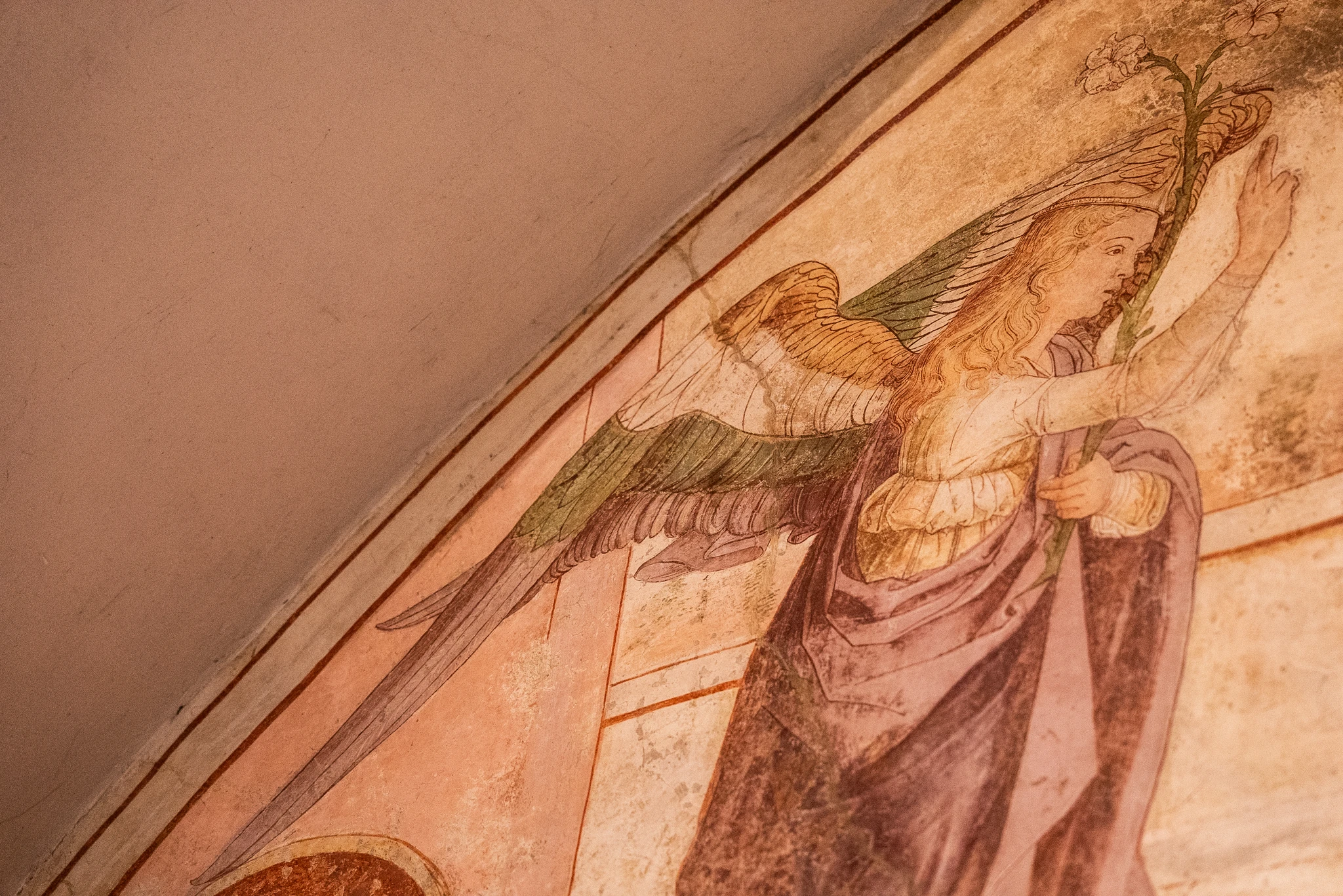 Affresco che raffigura una figura alata vista di profilo, con un braccio sollevato che regge un ramo fiorito. La figura indossa un abito lungo nei toni del viola e oro, e presenta grandi ali verdi e dorate. Lo sfondo è costituito da una superficie muraria dai toni chiari.