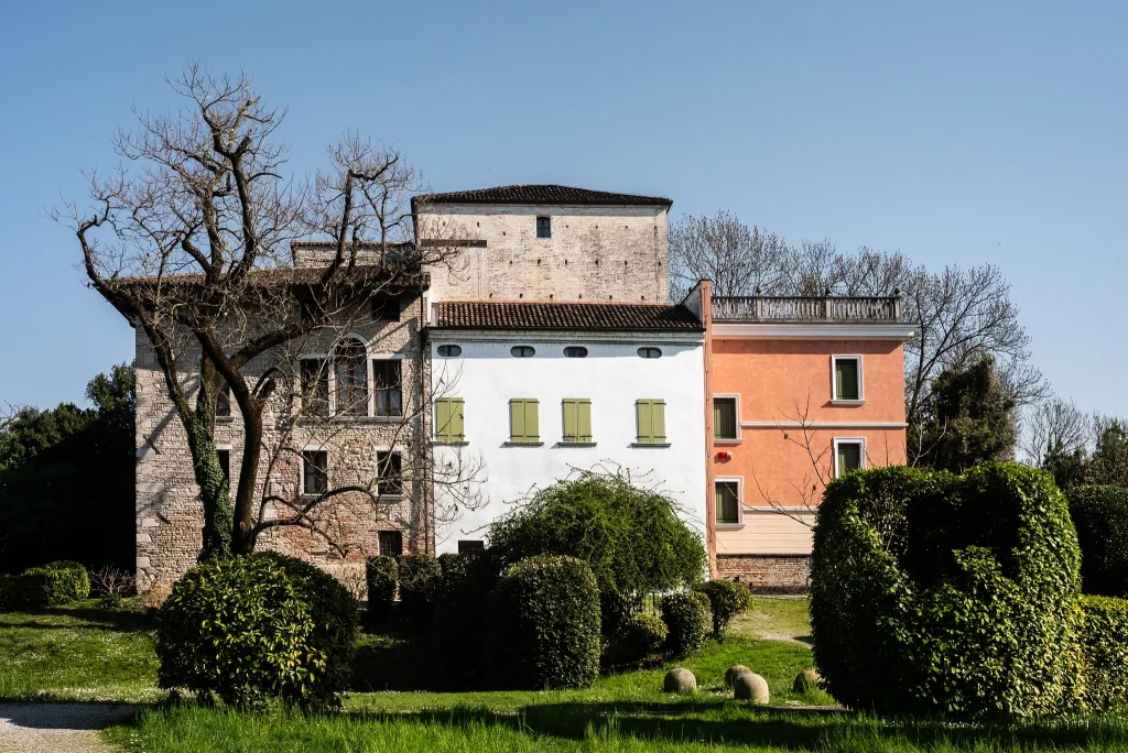 Edificio composto da tre corpi affiancati, con facciate di colori diversi: una struttura in mattoni a sinistra, una parte centrale bianca con imposte verdi e una sezione arancione a destra. Davanti all’edificio si trovano siepi potate e un prato verde. Sullo sfondo sono visibili alberi spogli e cielo sereno.