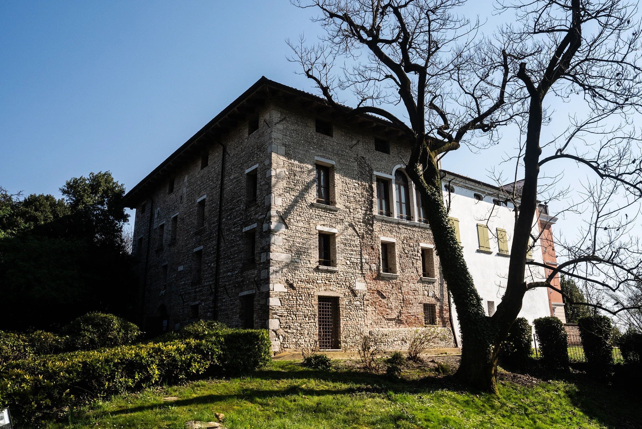 Edificio storico in pietra a più piani, con facciata irregolare e finestre di diverse dimensioni, situato su un terreno erboso. Accanto all’edificio si trova una struttura adiacente intonacata di bianco. In primo piano sono visibili cespugli potati e un albero spoglio dalle grandi ramificazioni.