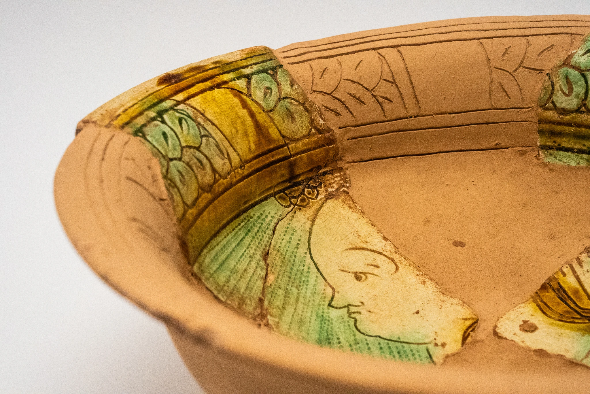 Ciotola in ceramica chiara con frammenti decorati reinseriti lungo il bordo interno. I frammenti presentano motivi verdi e gialli e parte del profilo di un volto stilizzato. Il resto della superficie interna è liscia e priva di decorazioni.