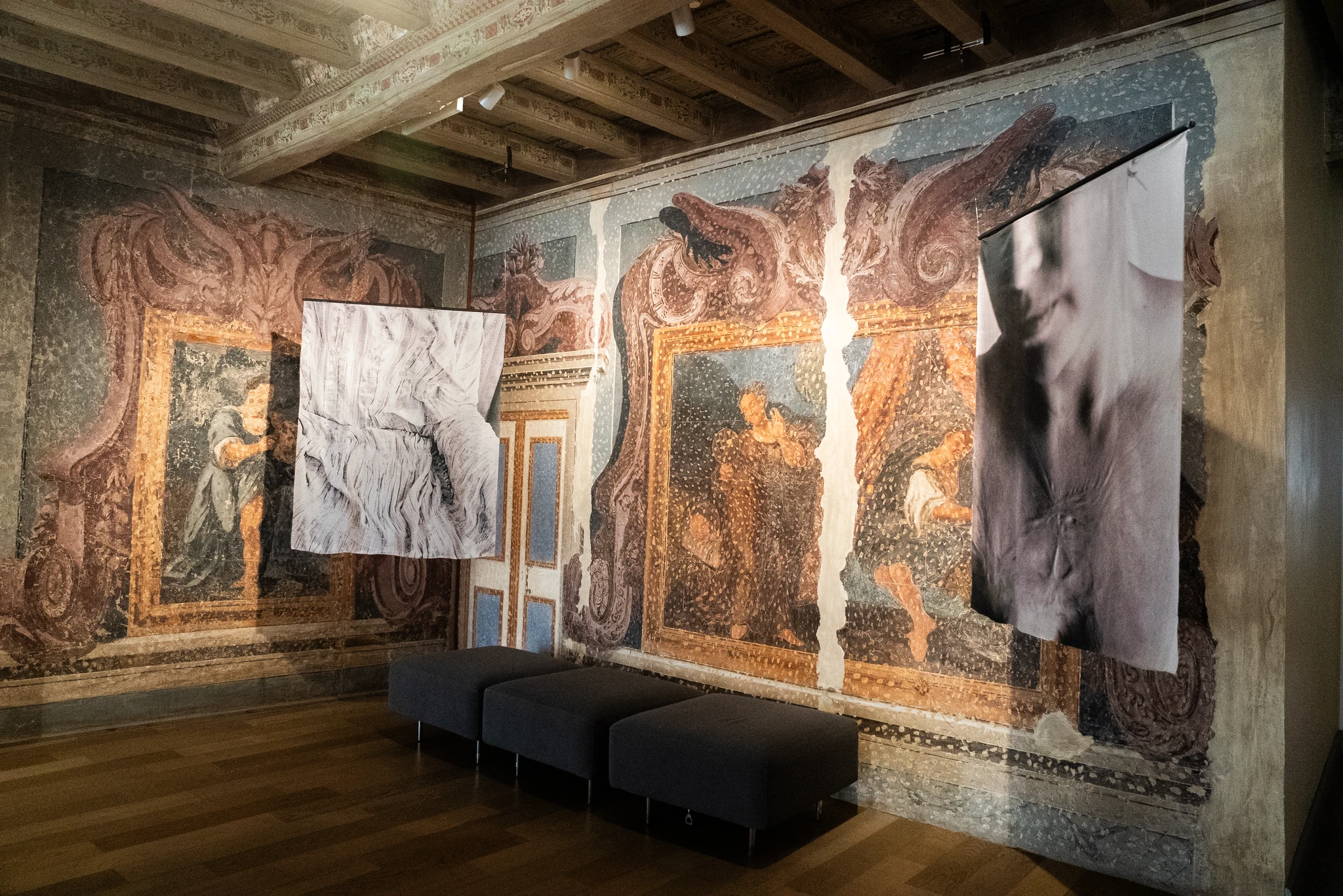 Interno di una sala con pareti decorate da grandi affreschi dai toni blu, arancioni e rosati, raffiguranti figure umane incorniciate da motivi ornamentali. Dal soffitto ligneo decorato scendono due grandi stampe fotografiche in bianco e nero, sospese al centro della stanza. Sotto le stampe sono collocati tre pouf scuri allineati. Il pavimento è in legno chiaro.