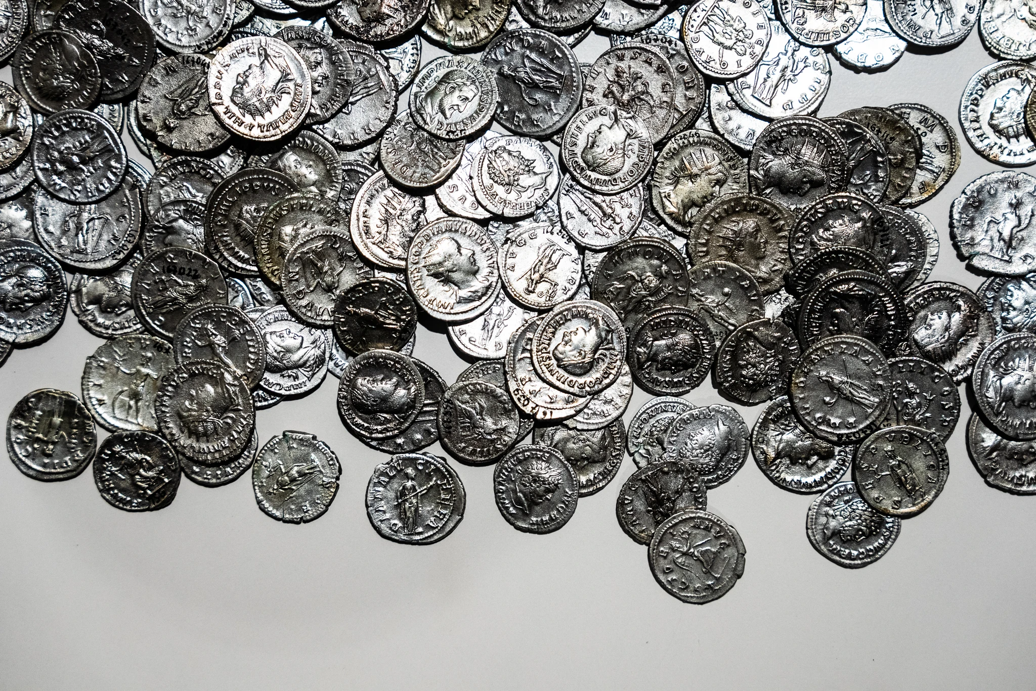 Ampio gruppo di monete metalliche antiche sparse su una superficie chiara. Le monete presentano rilievi con profili umani e iscrizioni lungo i bordi.