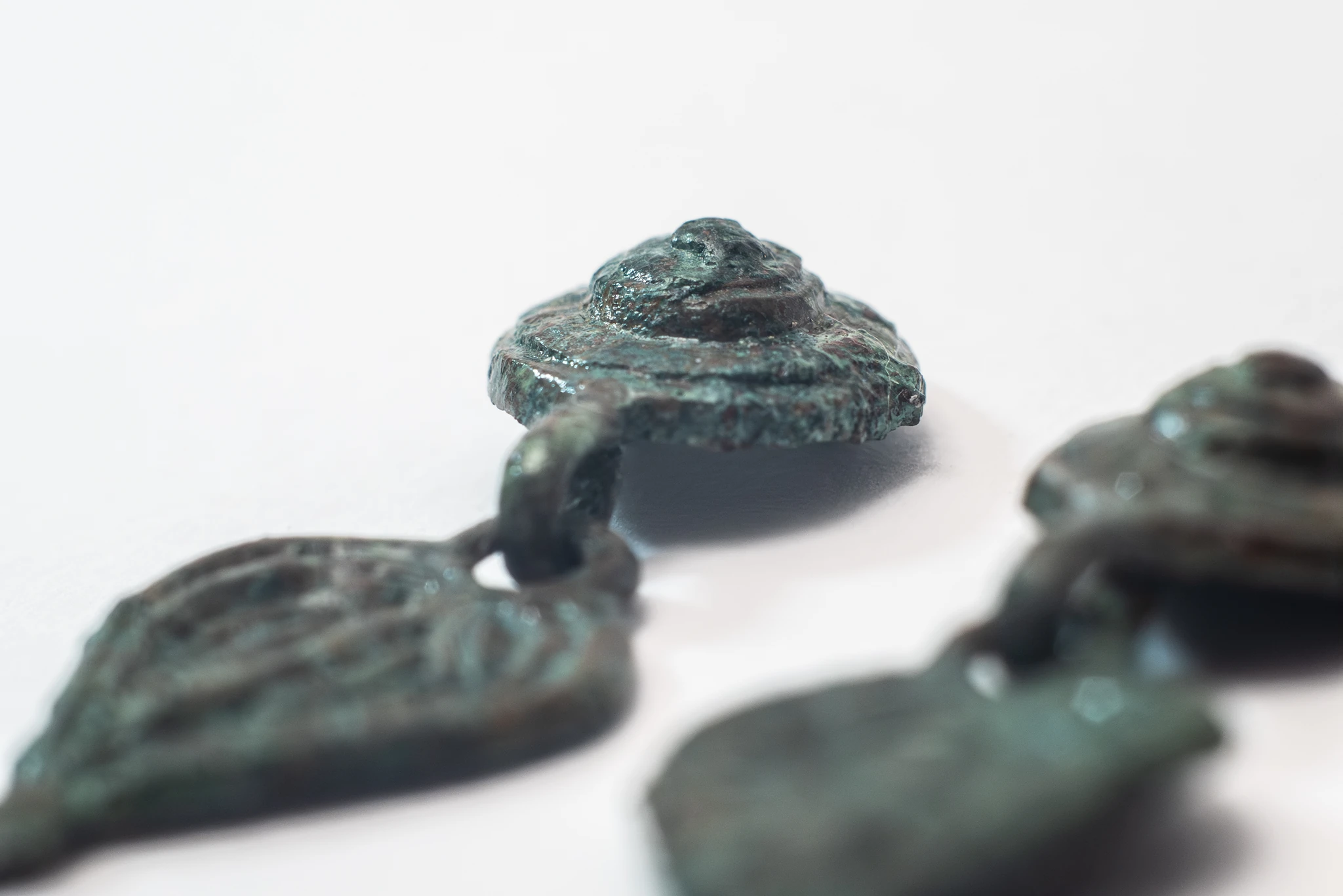 Primo piano di piccoli elementi metallici verdi ossidati, collegati tra loro da anelli. L’elemento centrale ha forma circolare e leggermente convessa, con superficie irregolare. Gli oggetti sono appoggiati su uno sfondo bianco, con alcune parti sfocate in primo piano e sullo sfondo.