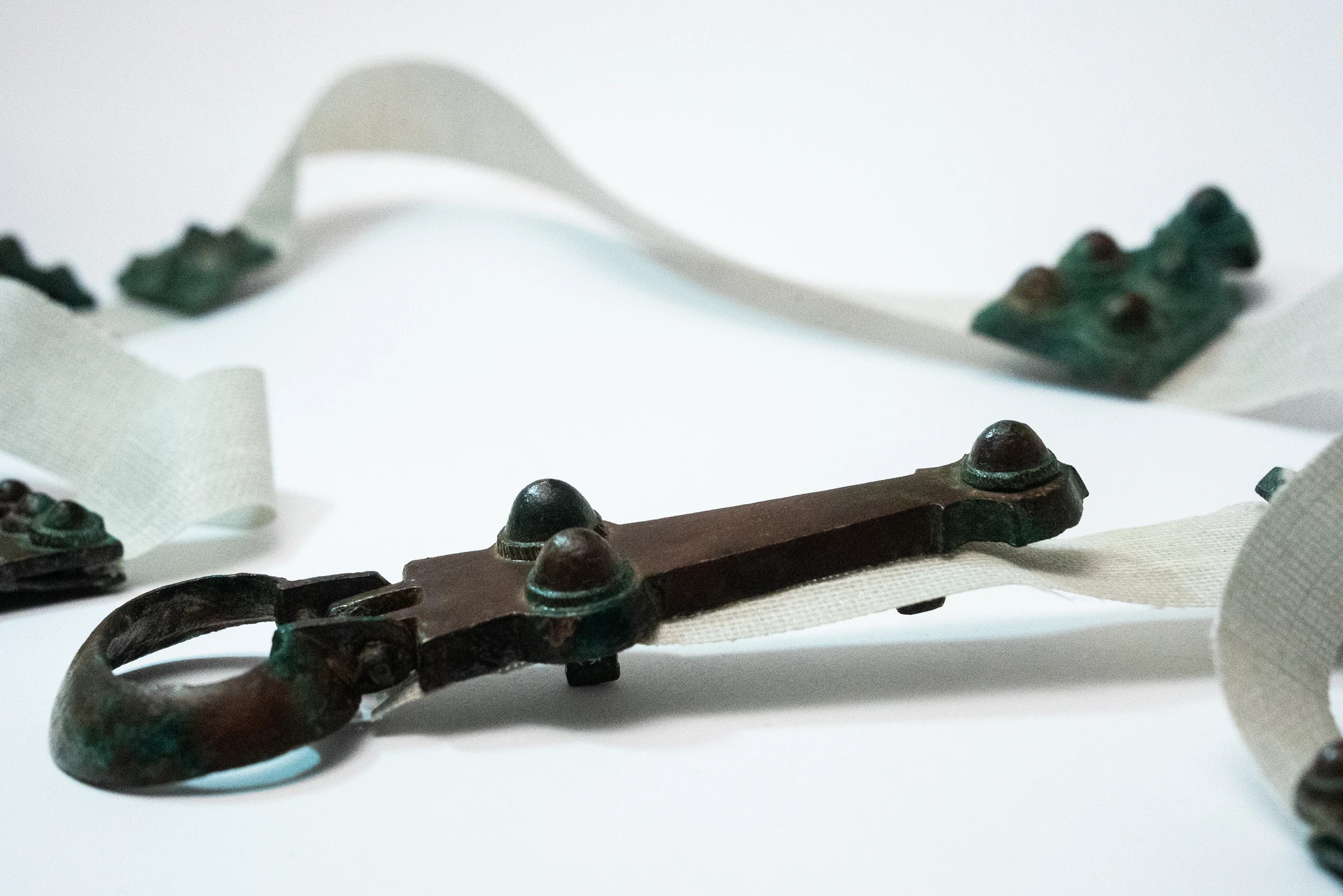 Primo piano di una fibbia metallica ossidata, di colore verde-bruno, fissata a una striscia tessile chiara. Sullo sfondo, sfocate, si intravedono altre placche metalliche e segmenti dello stesso nastro disposti sul piano.