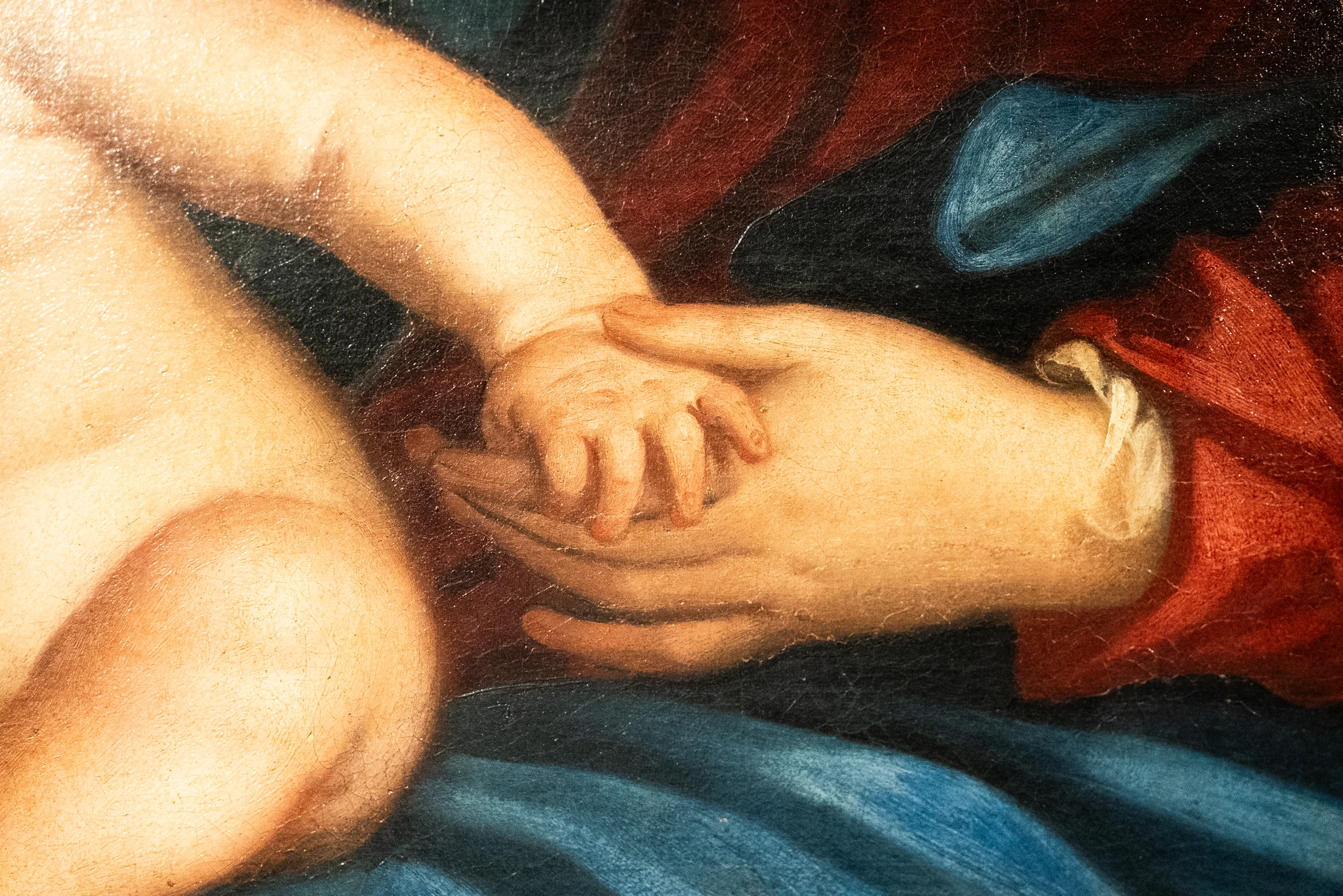 Dettaglio di un dipinto a olio che ritrae la mano di un adulto che sorregge con dolcezza la mano piccola di un neonato. Sullo sfondo si notano panneggi di vesti nei colori rosso e blu.