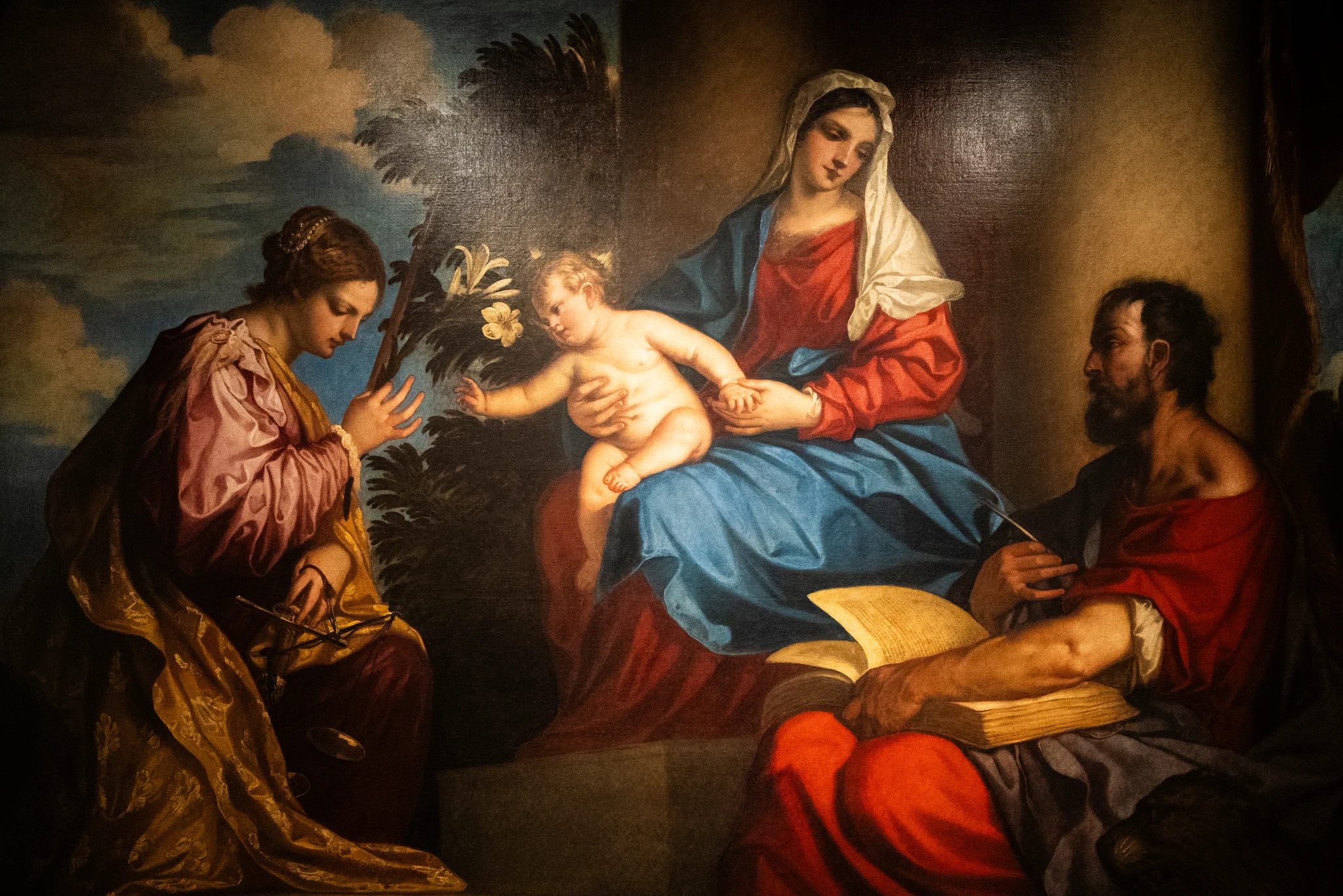 Dipinto che raffigura una scena religiosa: al centro una figura seduta con un bambino in braccio, entrambi con vesti dai toni del rosso e del blu. Alla sinistra della scena è presente una figura inginocchiata che regge un oggetto con forma circolare e fiori bianchi. A destra una figura seduta tiene aperto un grande libro. Lo sfondo mostra colonne architettoniche e un cielo con nuvole.