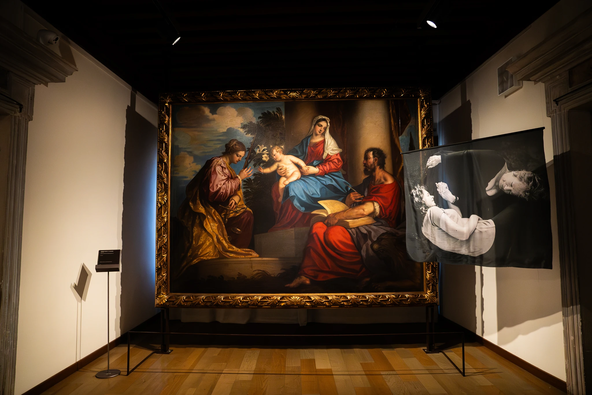 Sala espositiva con un grande dipinto a tema religioso incorniciato in oro al centro della parete. L’opera mostra più figure sedute e una figura infantile al centro della composizione. A destra del dipinto è esposto un pannello trasparente con una stampa in bianco e nero raffigurante una figura distesa. Il pavimento è in legno e l’illuminazione proviene da spot sul soffitto.