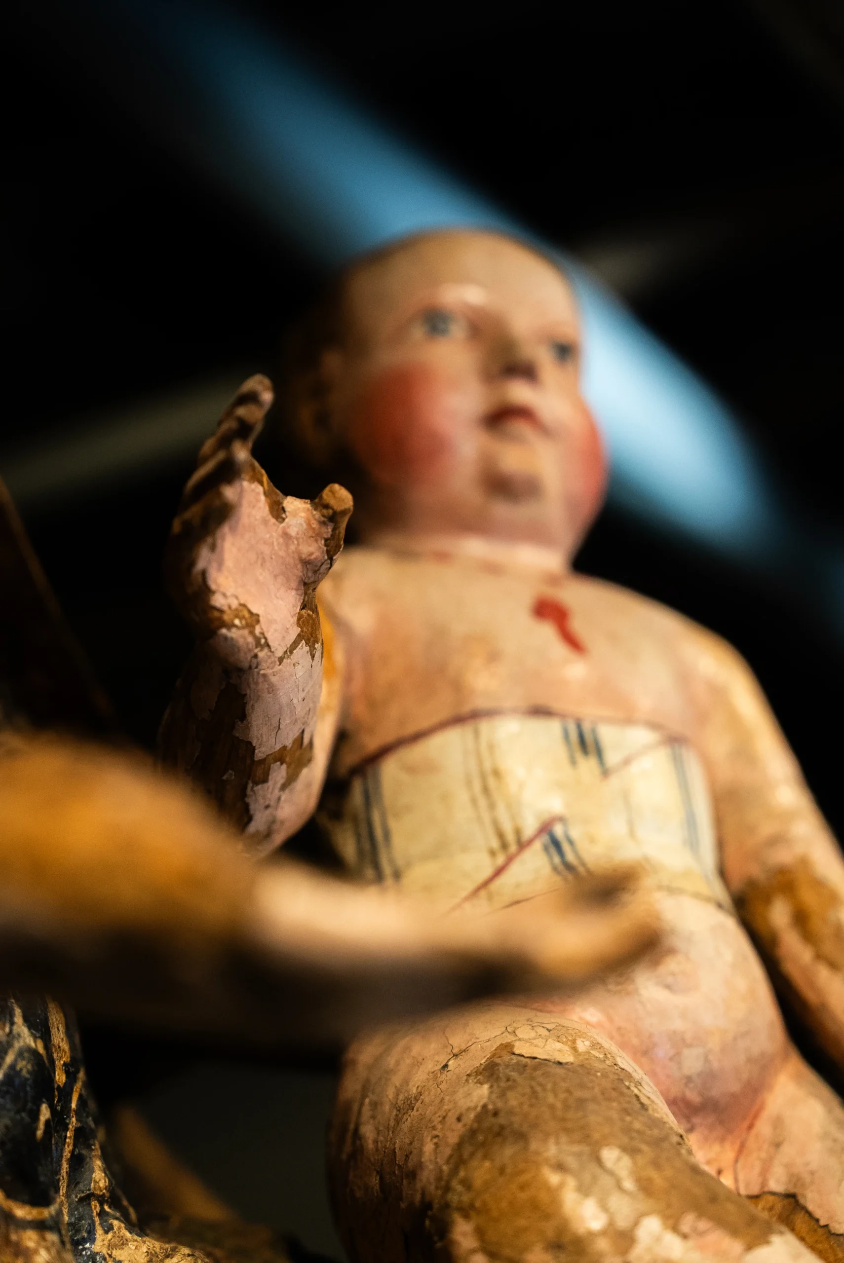 Primo piano di una scultura lignea dipinta raffigurante una figura infantile seduta. La superficie presenta ampie aree di usura con la pittura scrostata. La mano in primo piano è sollevata e mostra crepe nella materia. Sul busto è visibile una fascia dipinta con linee verticali e un piccolo segno rosso al centro. Lo sfondo è scuro e sfocato.