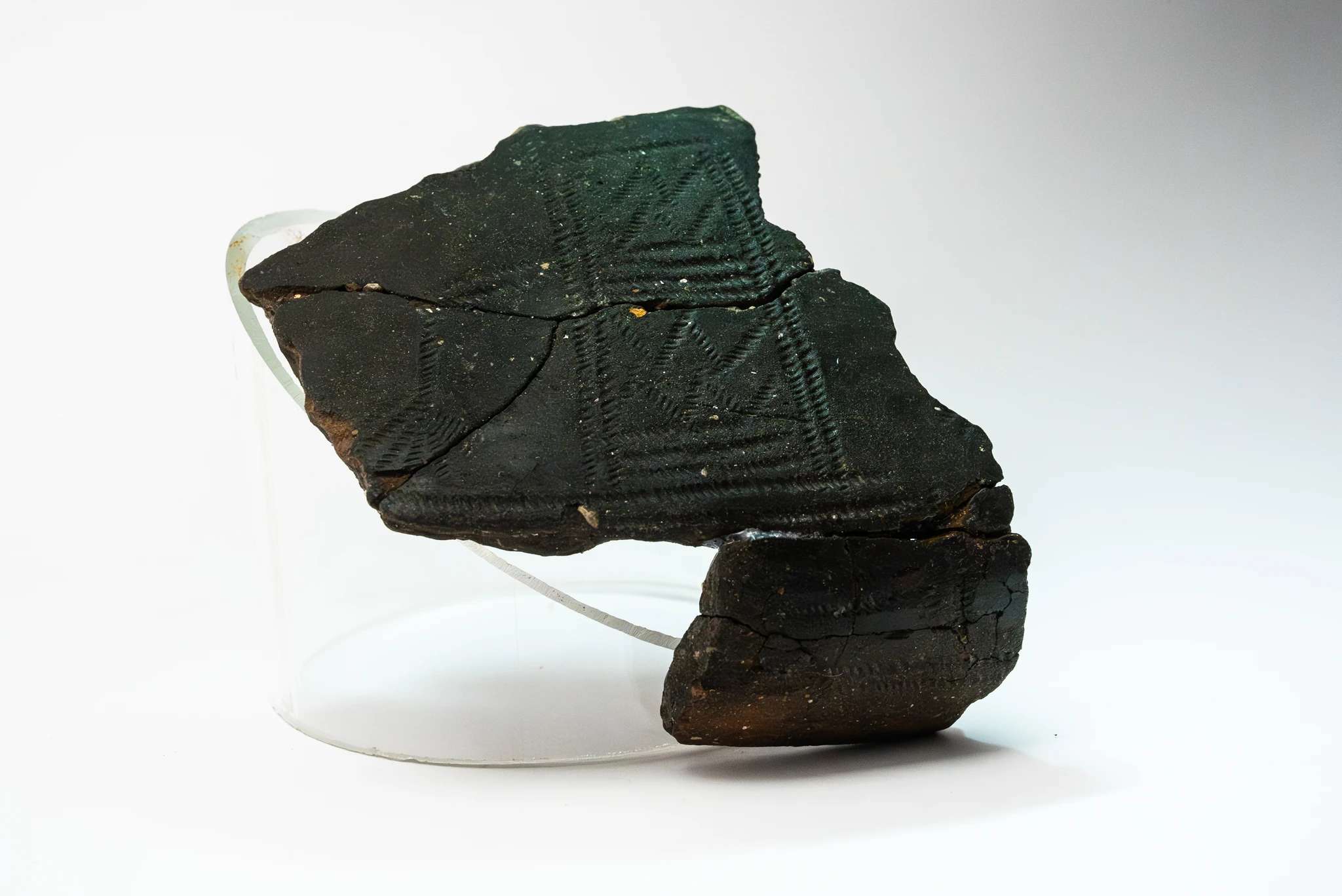 Frammenti ricomposti di un manufatto ceramico scuro, decorato con motivi geometrici incisi. I pezzi, uniti in una forma incompleta, sono esposti su un supporto trasparente contro uno sfondo bianco.