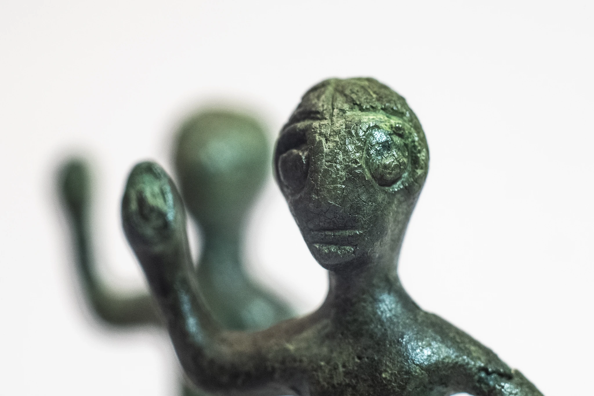 Primo piano di una piccola statuetta antropomorfa in metallo di colore verde, con superficie irregolare e tratti del volto incisi. La figura ha un braccio sollevato. Sullo sfondo, sfocata, è visibile un’altra statuetta simile.