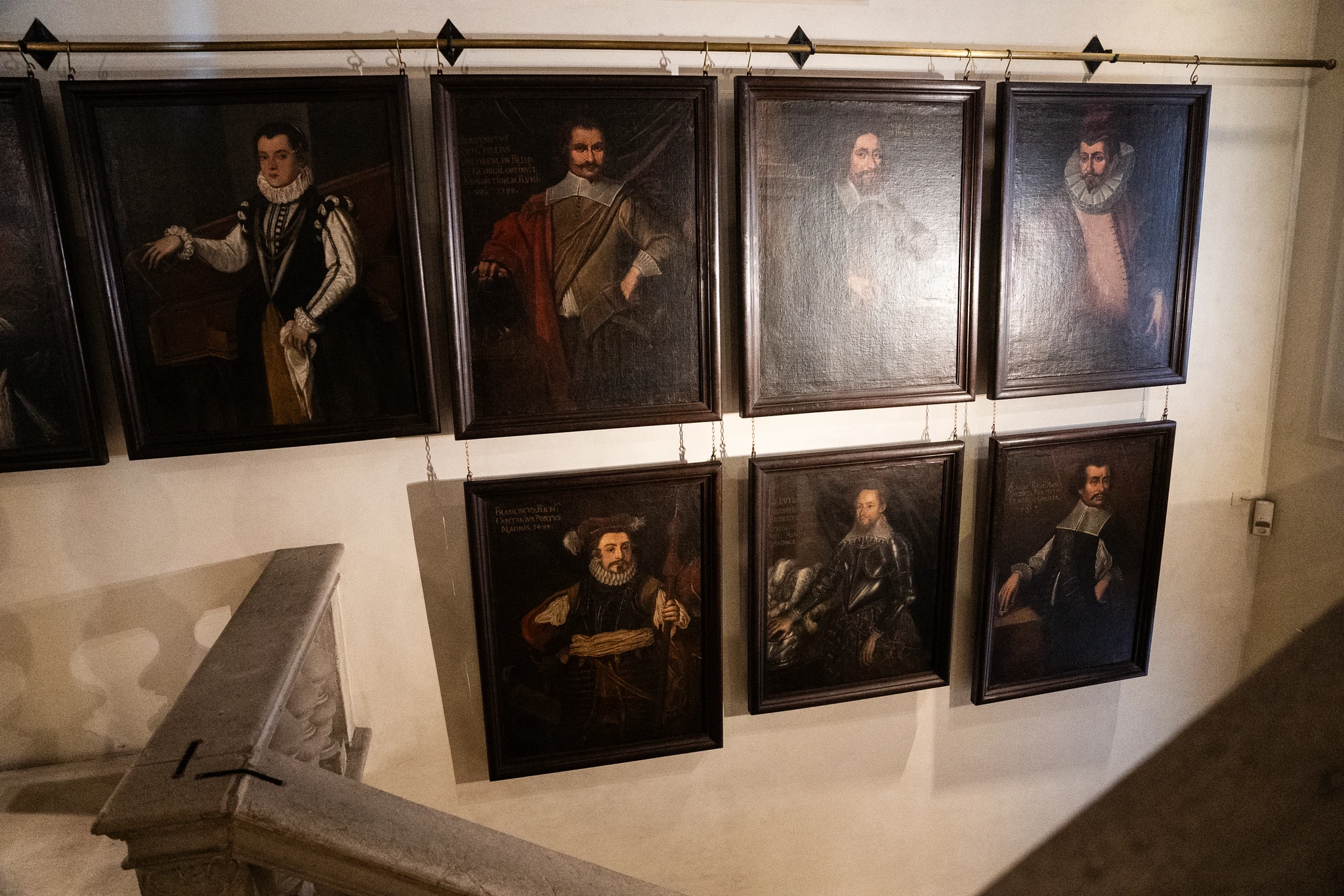 Parete interna di un edificio con otto ritratti dipinti a olio, ciascuno in cornice scura, disposti su due file. I dipinti raffigurano figure vestite con abiti storici. In basso a sinistra è visibile una parte della scala in pietra con corrimano scolpito.