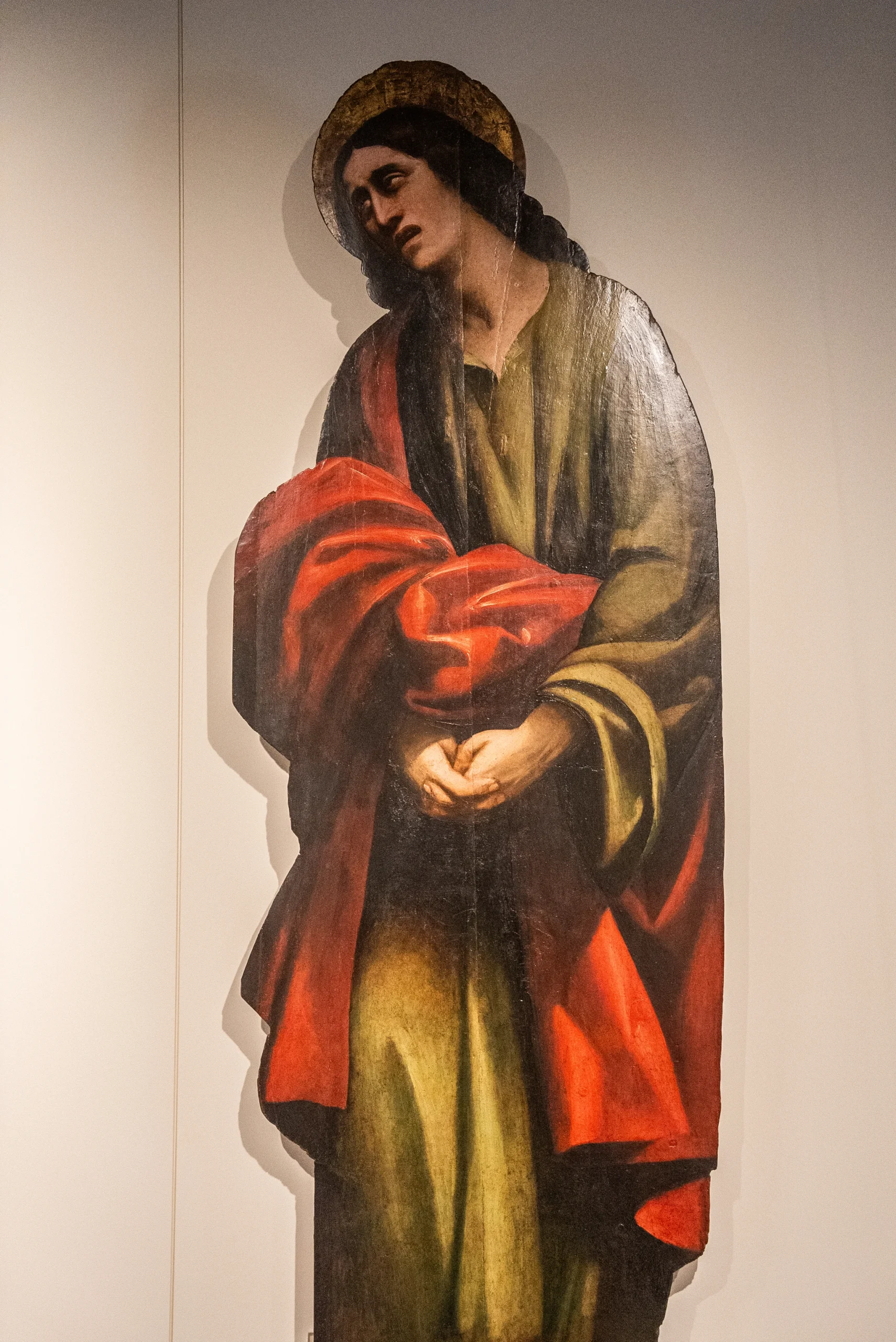 Riproduzione fotografica di una figura dipinta a grandezza quasi naturale su un pannello verticale. La figura è rappresentata in posizione eretta, con il capo leggermente inclinato verso un lato e le mani unite davanti al corpo. Indossa un abito lungo dai toni scuri con drappeggi e un ampio mantello rosso avvolto attorno alle braccia. Sul capo è presente un’aureola di colore dorato. Lo sfondo è semplice e uniforme, illuminato da una luce diretta che evidenzia la superficie pittorica.