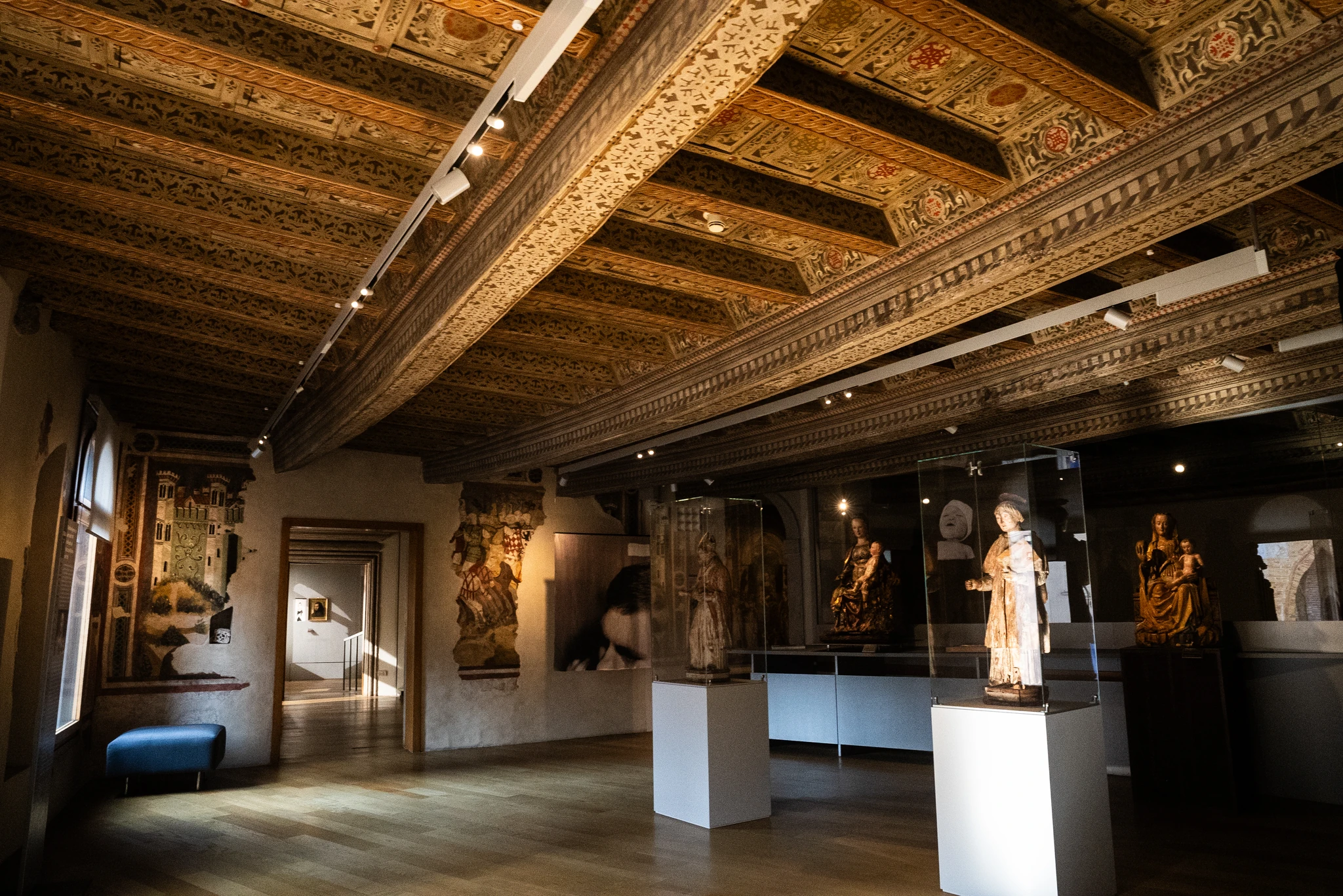 Interno di una sala museale con soffitto ligneo decorato da motivi geometrici e floreali. La stanza espone diverse sculture in teche trasparenti su piedistalli bianchi e altre opere lungo le pareti. Sulla sinistra si vede una finestra e un corridoio che conduce a un altro ambiente illuminato. Il pavimento è in legno chiaro.
