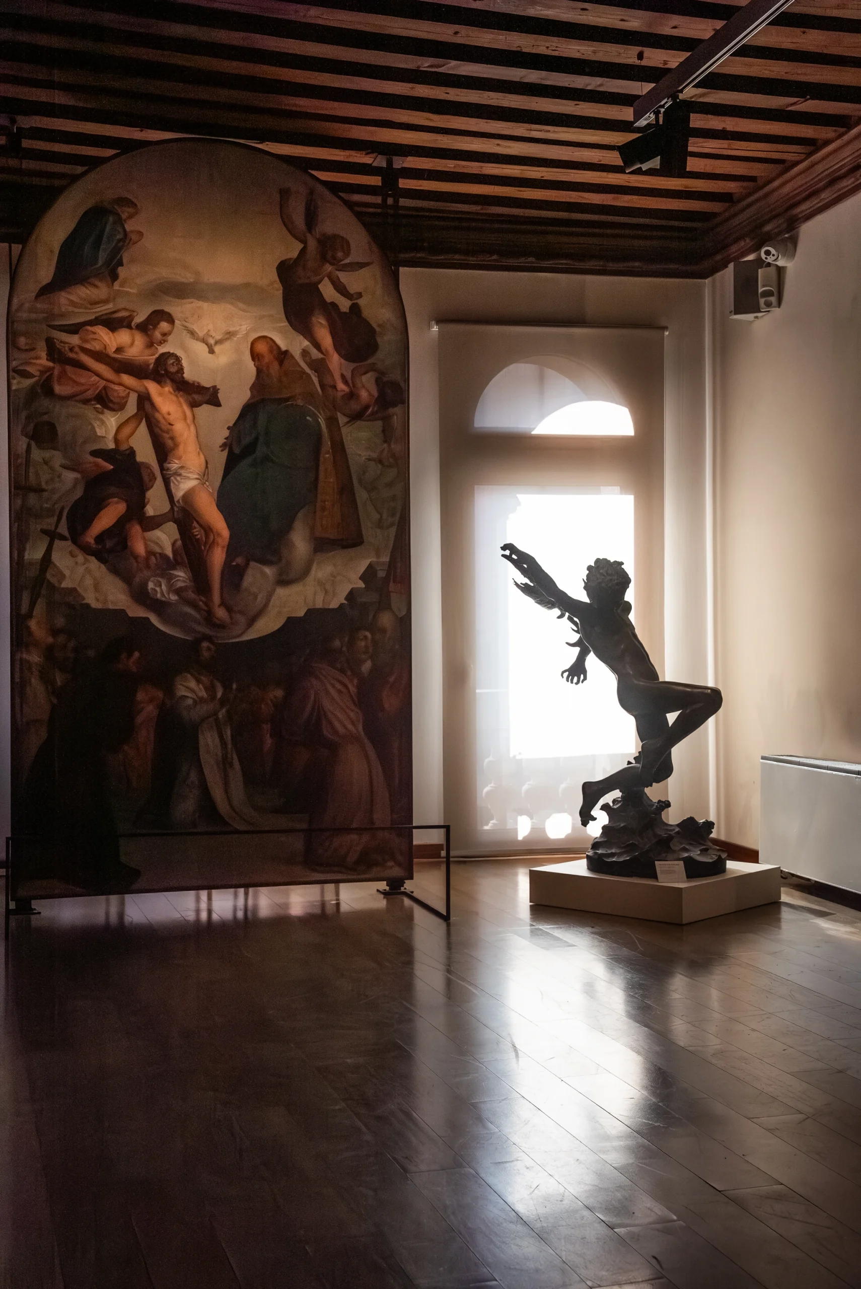 Interno di una sala espositiva con un grande dipinto religioso su pannello a sinistra e una scultura scura su piedistallo a destra. La scultura è illuminata dalla luce proveniente da una porta-finestra sullo sfondo, creando una silhouette marcata. Il pavimento è in legno e il soffitto presenta travi a vista.
