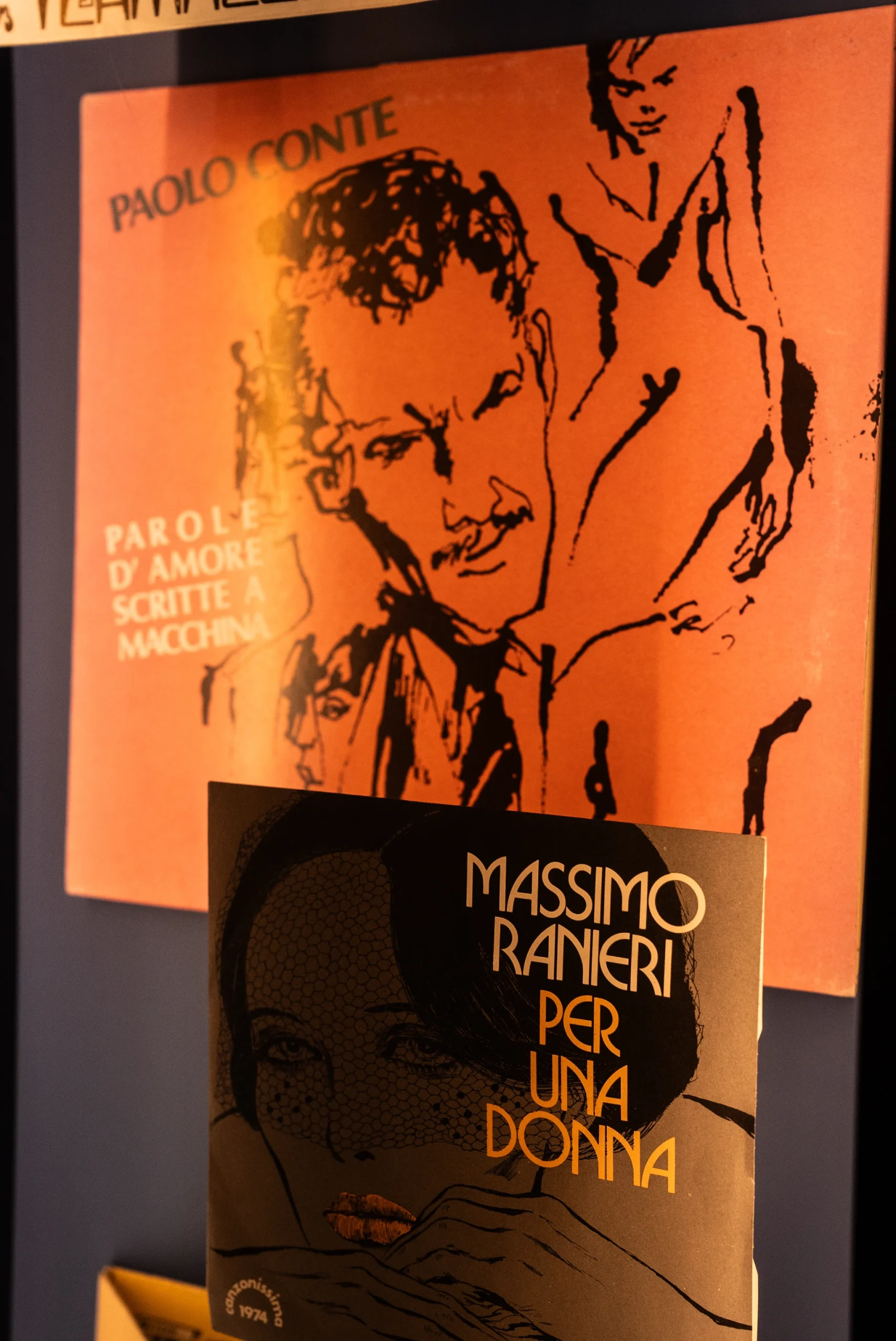Due copertine di dischi esposte verticalmente: sullo sfondo una copertina arancione con illustrazione lineare di un volto e figura, accompagnata dal testo ‘Paolo Conte’ e da altre scritte. In primo piano una copertina più piccola nei toni del nero e ocra con un’illustrazione stilizzata di un volto e la scritta ‘Massimo Ranieri – Per una donna’.