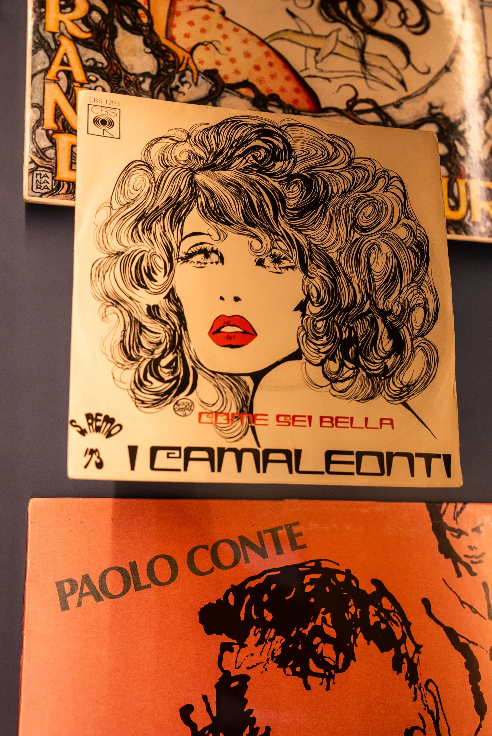 Copertina di un disco con illustrazione in bianco e nero di una figura con capelli voluminosi e labbra rosse. In alto è visibile la scritta ‘COME SEI BELLA’ e in basso ‘I CAMALEONTI’. Dietro la copertina, parzialmente visibile, c’è un’altra copertina con illustrazione colorata. Nella parte bassa dell’immagine appare anche una copertina arancione con il nome ‘Paolo Conte’.