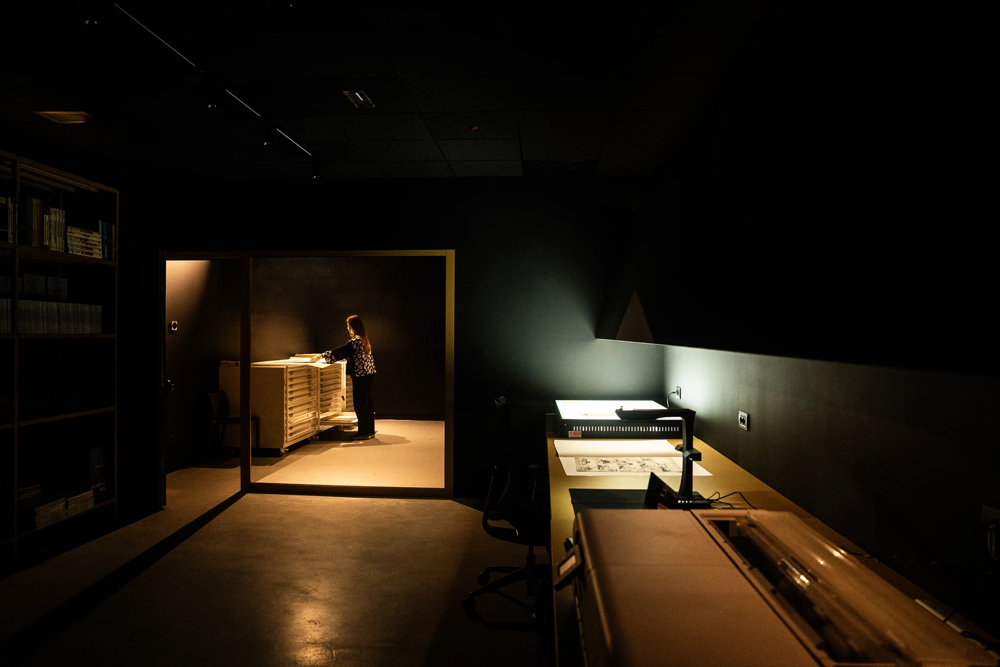 In un'immagine fotografica scattata all'interno del caveau del museo del fumetto di pordenone, in una stanza oscura, si scorgono due zone principali, separate da un divisorio in vetro che appare come una vetrina o una finestra. La zona interna, più chiara, mostra un armadio o una serie di cassettiere dove si possono scorgere stampe e fumetti organizzati in pile ordinate. All'interno si trova una persona che sta esaminando uno di questi cassetti. La zona esterna, più scura e immersa in un'illuminazione a basso livello, presenta una scrivania con un monitor e alcuni fogli sparsi che mostrano tavole di fumetti. A sinistra si può notare una libreria che ospita volumi di fumetti.