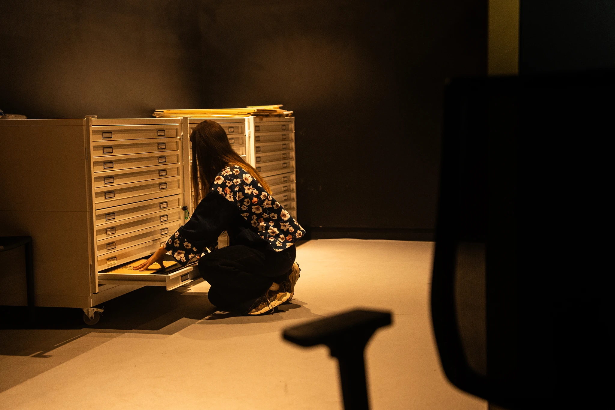 Ambiente interno con illuminazione soffusa. Una persona è accovacciata davanti a una grande cassettiera a cassetti piatti, uno dei quali è aperto. La persona sta consultando o sistemando dei fogli o documenti all’interno del cassetto. In primo piano, sul lato destro, è visibile la sagoma sfocata di uno schienale di sedia.