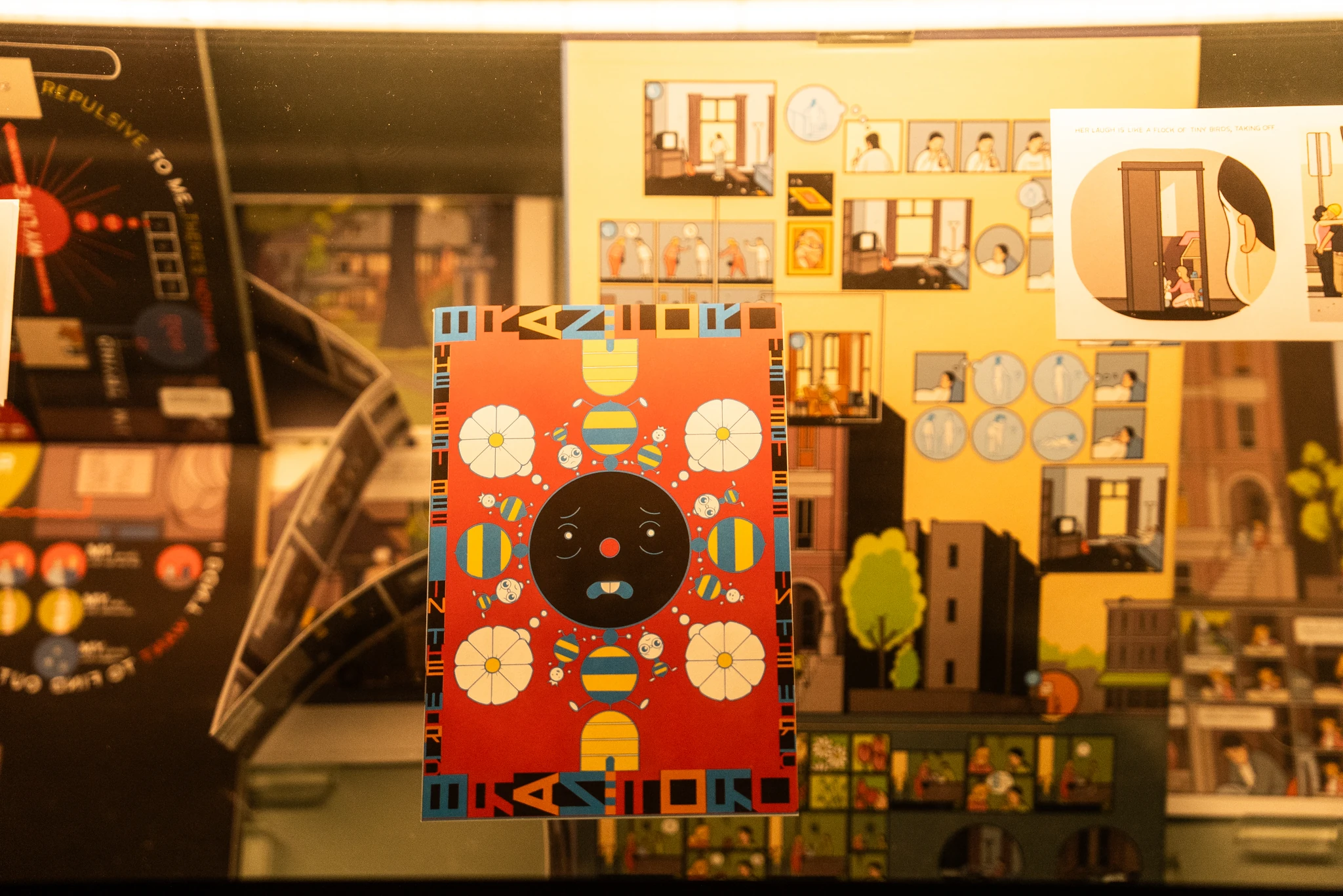 Vetrina con diversi pannelli illustrati dai colori vivaci. Al centro è esposta un’immagine rettangolare su sfondo rosso, con un elemento decorativo circolare nero attorniato da forme geometriche colorate. Attorno, su pannelli gialli e scuri, compaiono vignette e disegni che rappresentano interni di edifici, figure stilizzate e scene a fumetti.