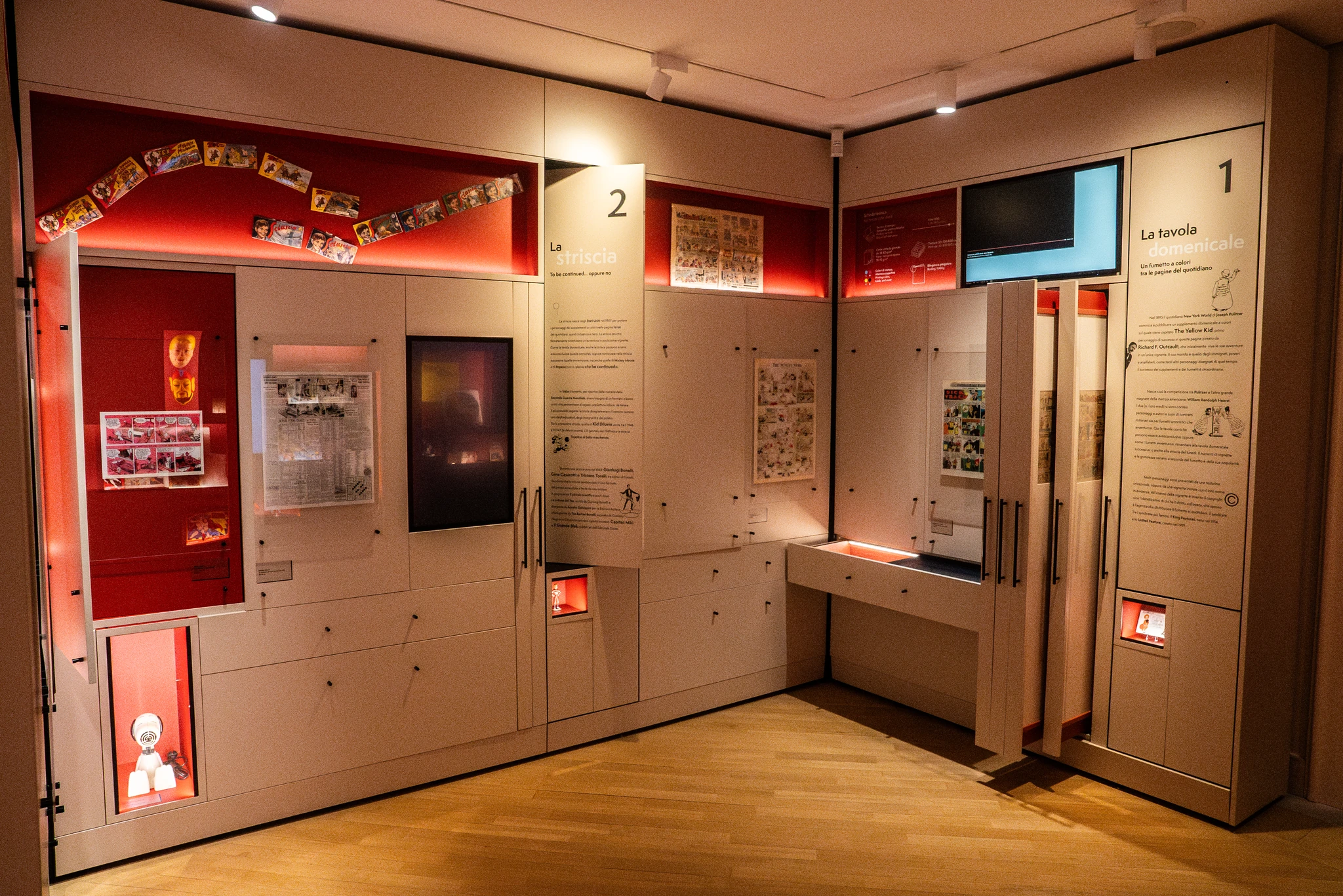 Vetrina museale composta da moduli espositivi chiari con pannelli rossi e superfici retroilluminate. Sono visibili fumetti, illustrazioni e piccoli oggetti distribuiti in teche verticali e orizzontali. I pannelli numerati ‘1’ e ‘2’ riportano testi informativi. Il pavimento è in legno chiaro e l’area è illuminata da luci a soffitto.