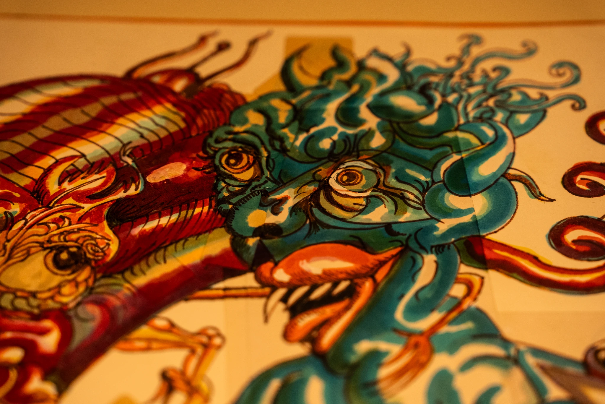 Dettaglio ravvicinato di un’illustrazione dai colori vivaci che raffigura una figura fantastica con pelle blu, occhi grandi e linee sinuose che si estendono dalla testa. Accanto alla figura è presente un altro elemento illustrato in tonalità rosse e gialle, con forme organiche e tratteggi marcati.