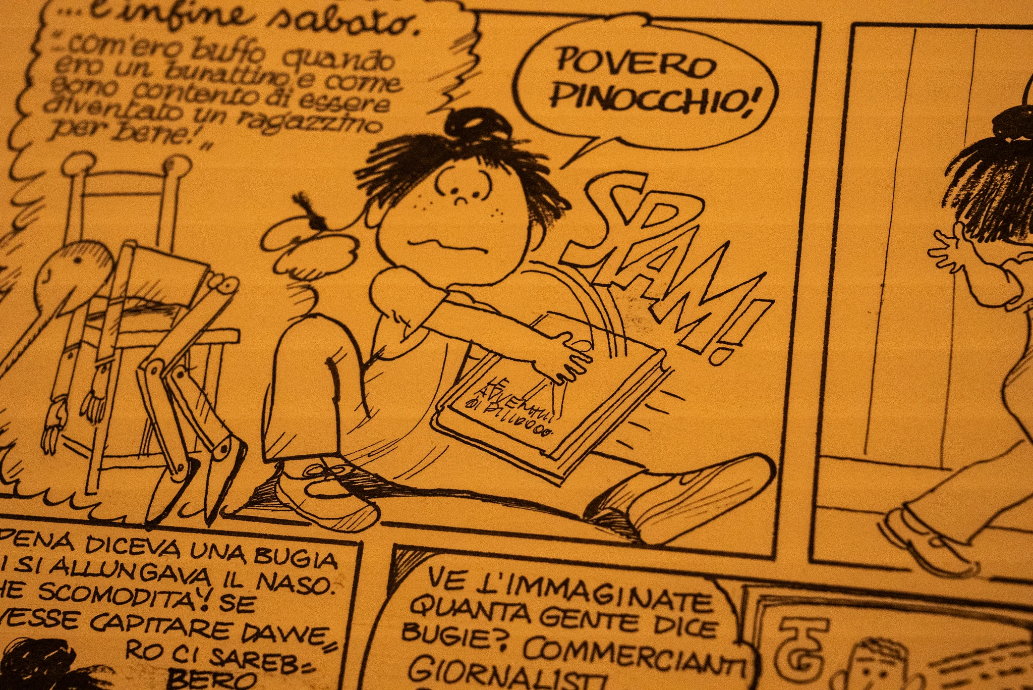 Vignetta di un fumetto disegnata con linee nere su sfondo giallo. Al centro è presente una figura seduta a terra con un libro tra le mani; accanto al libro compare la scritta 'SPAM'. Sopra la figura, in un fumetto, è riportato il testo 'Povero Pinocchio!'. A sinistra si vede un manichino articolato appoggiato a una sedia, mentre nella parte superiore e inferiore della scena compaiono altri riquadri con testo in italiano.