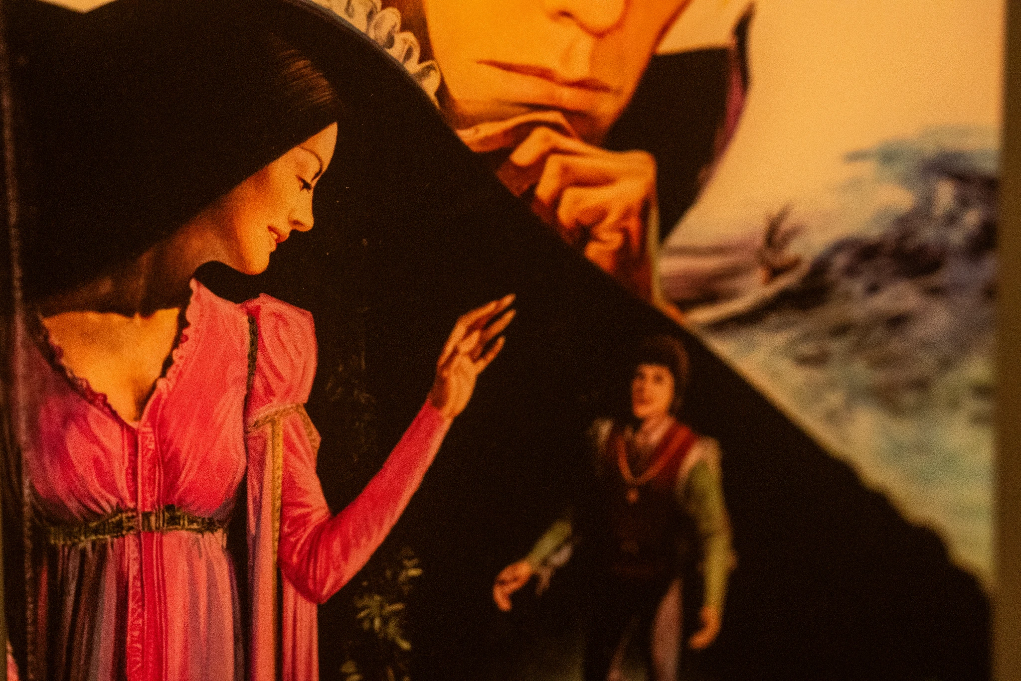 Dettaglio di un’illustrazione che raffigura una figura in primo piano con abito rosa e braccio sollevato. Sullo sfondo si vede una figura più piccola in abiti scuri e, in alto, una parte del volto di un’altra figura con mano appoggiata al mento. Sul lato destro sono presenti forme pittoriche dai toni blu e bianchi.