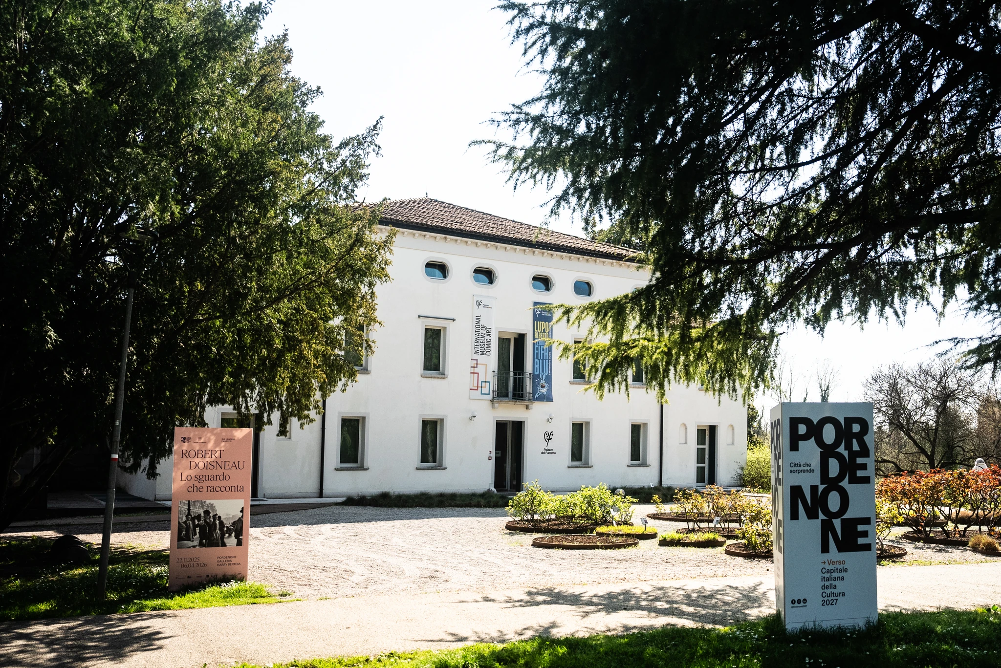 Edificio bianco a due piani con finestre regolari e un ingresso centrale, circondato da alberi e aiuole. Davanti all’edificio sono presenti due pannelli informativi verticali, uno rosa a sinistra e uno bianco a destra con la scritta ‘Pordenone’. Il cielo è luminoso e l’area è immersa in un ambiente verde.