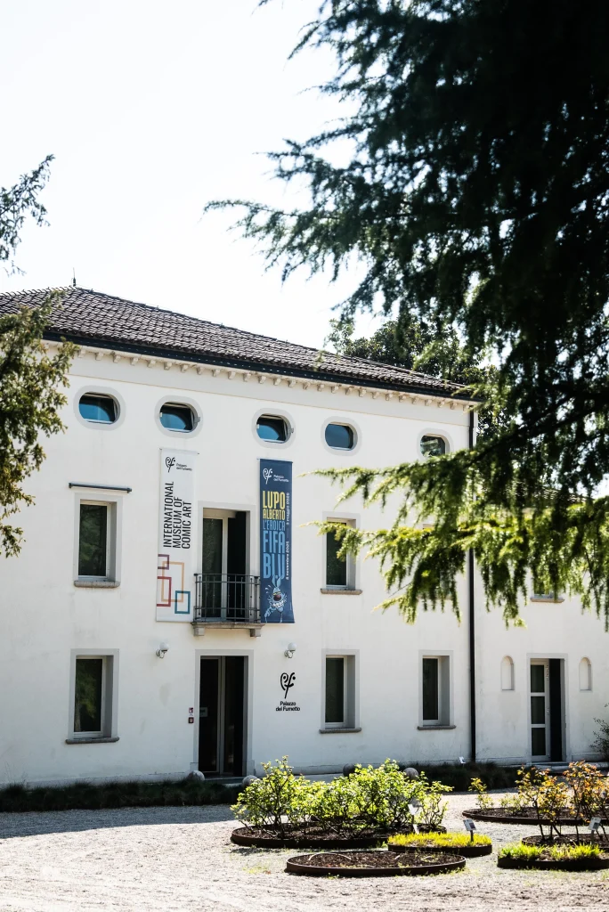 Edificio bianco a due piani con finestre ovali al piano superiore e un ingresso centrale. Sulla facciata sono appesi due banner verticali con testo e grafiche. Davanti all’edificio si trovano aiuole verdi e una zona di ghiaia, mentre rami di alberi incorniciano parzialmente la vista.