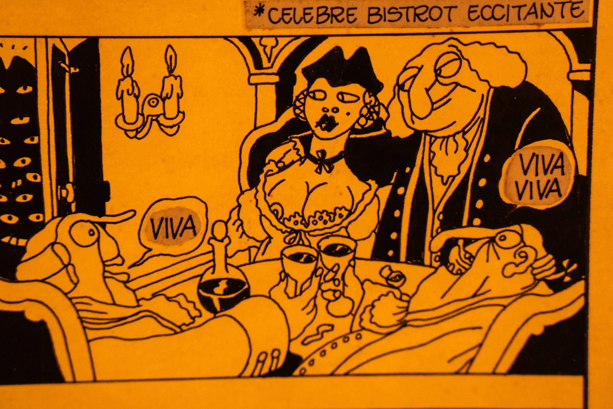 Vignetta illustrata dai toni giallo-arancio che mostra quattro figure stilizzate sedute attorno a un tavolo imbandito con bicchieri e una bottiglia. Due figure al centro sono vicine tra loro, una indossa un cappello scuro e un abito decorato. Ai lati del tavolo sono presenti due figure reclinate con fumetti contenenti la parola ‘VIVA’. Sullo sfondo si vedono elementi architettonici stilizzati e due candelabri.