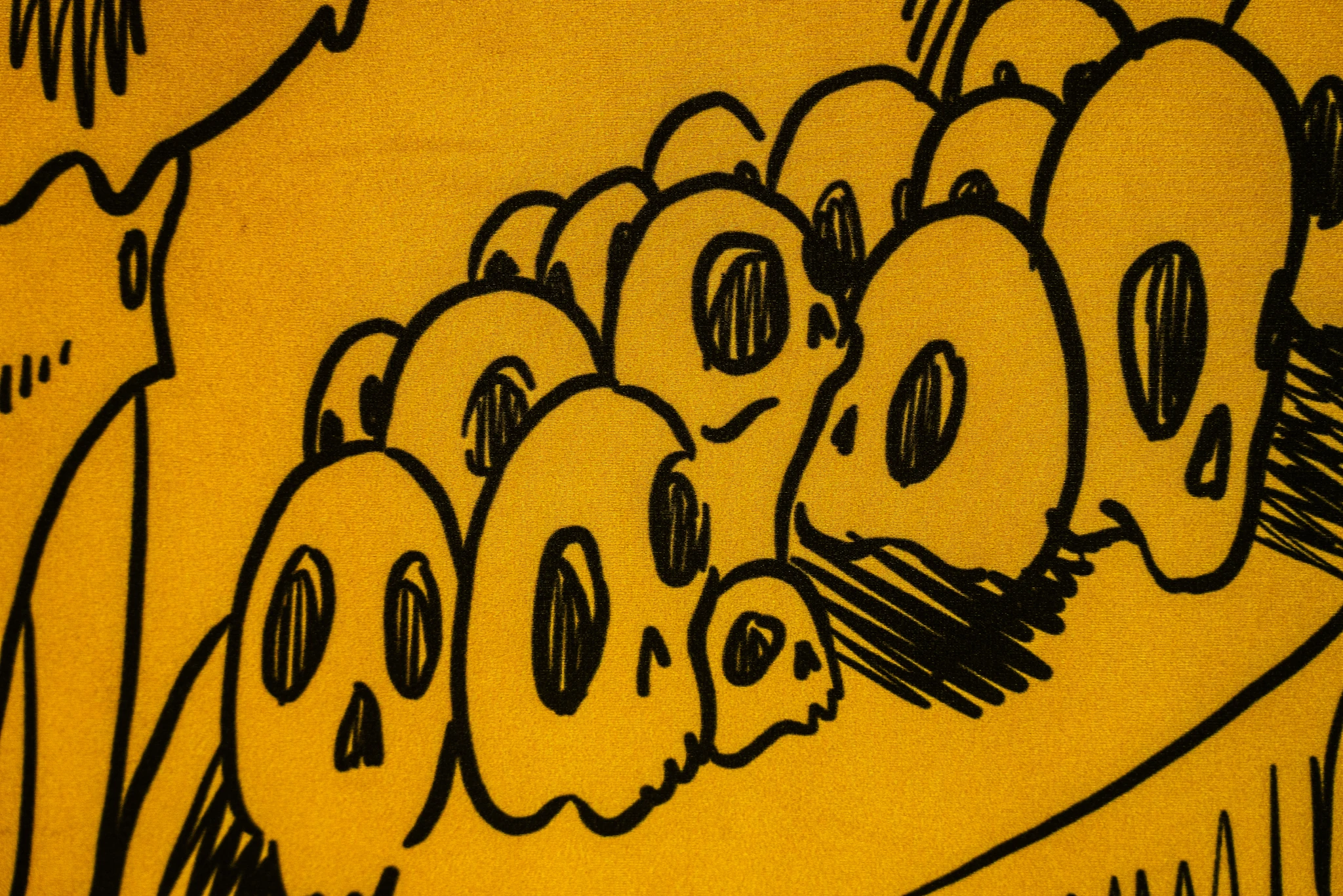 Un dettaglio di un'illustrazione grafica che raffigura un ammasso di teschi stilizzati. I teschi sono disegnati con tratti neri spessi ed essenziali su uno sfondo giallo ocra vibrante. Le orbite oculari e le cavità nasali sono rese con semplici fori ovali neri o tratteggiati.