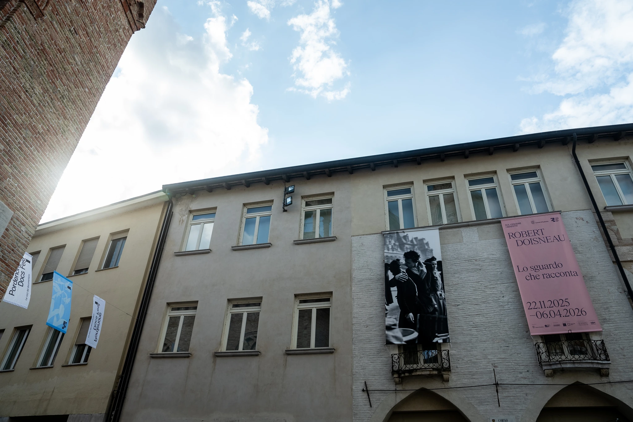 Facciata di un edificio a più piani con finestre rettangolari e due grandi banner verticali appesi: uno con una fotografia in bianco e nero e uno rosa con testo stampato. Accanto, sulla sinistra, sono appese altre bandiere verticali. In alto si vede una porzione di cielo con nuvole.