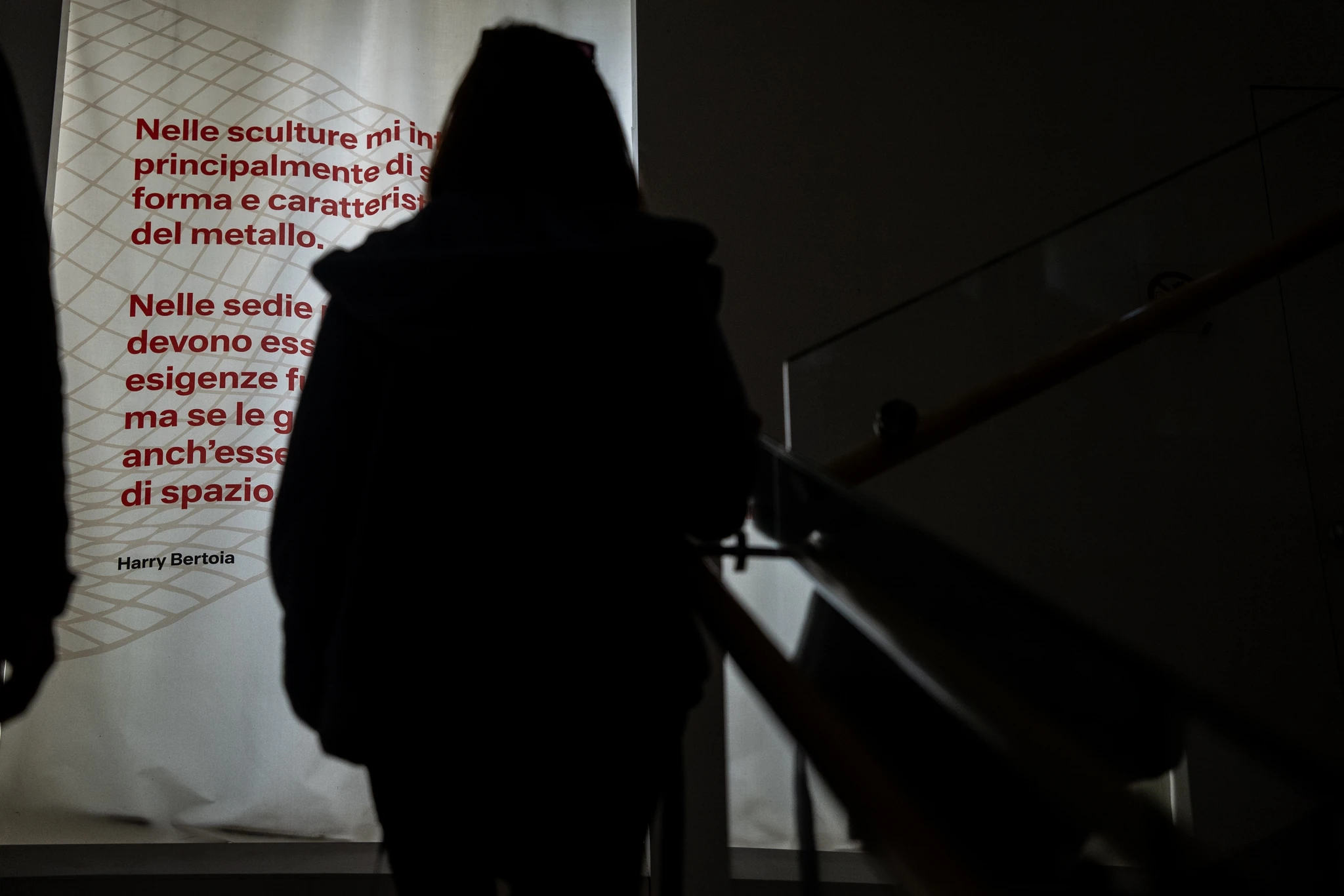 Silhouette di una persona in controluce che sale una scala interna, vista di spalle. Davanti alla persona è visibile un grande pannello illuminato con testo rosso su sfondo chiaro. A destra si intravede la ringhiera della scala.