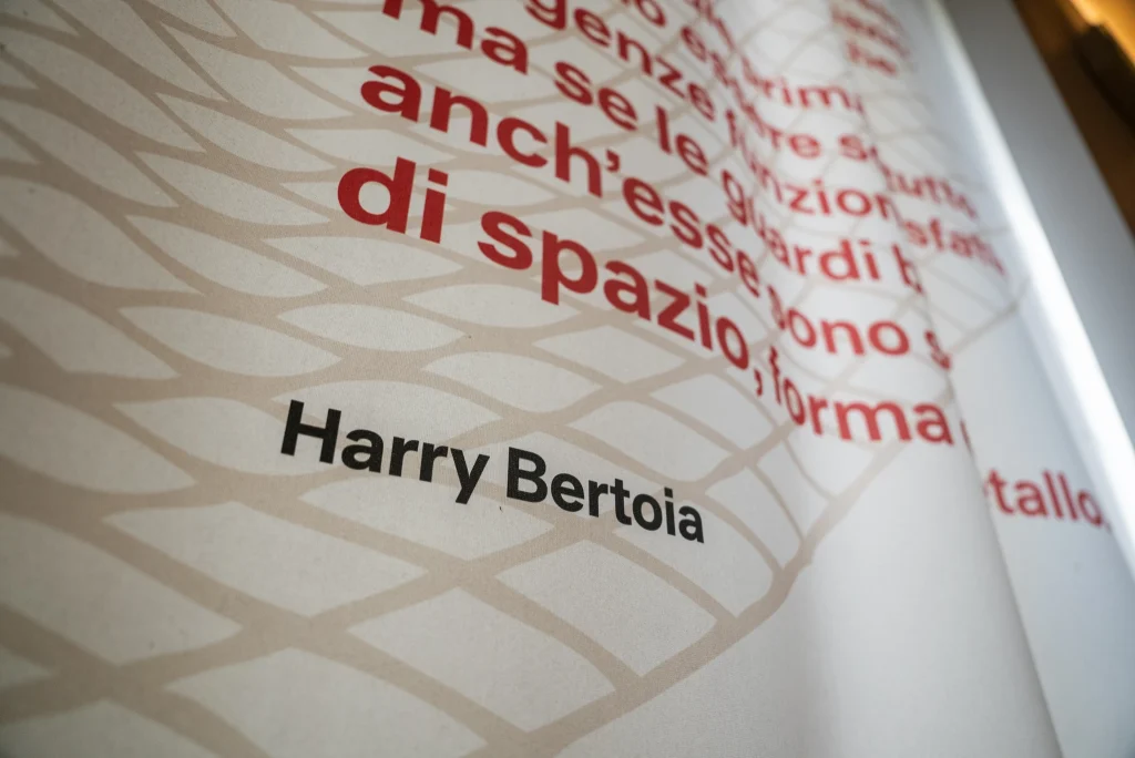 Dettaglio ravvicinato di un pannello espositivo, stampato con motivo geometrico beige sullo sfondo e testo rosso di grandi dimensioni. In basso al centro compare la scritta ‘Harry Bertoia’ in caratteri neri.