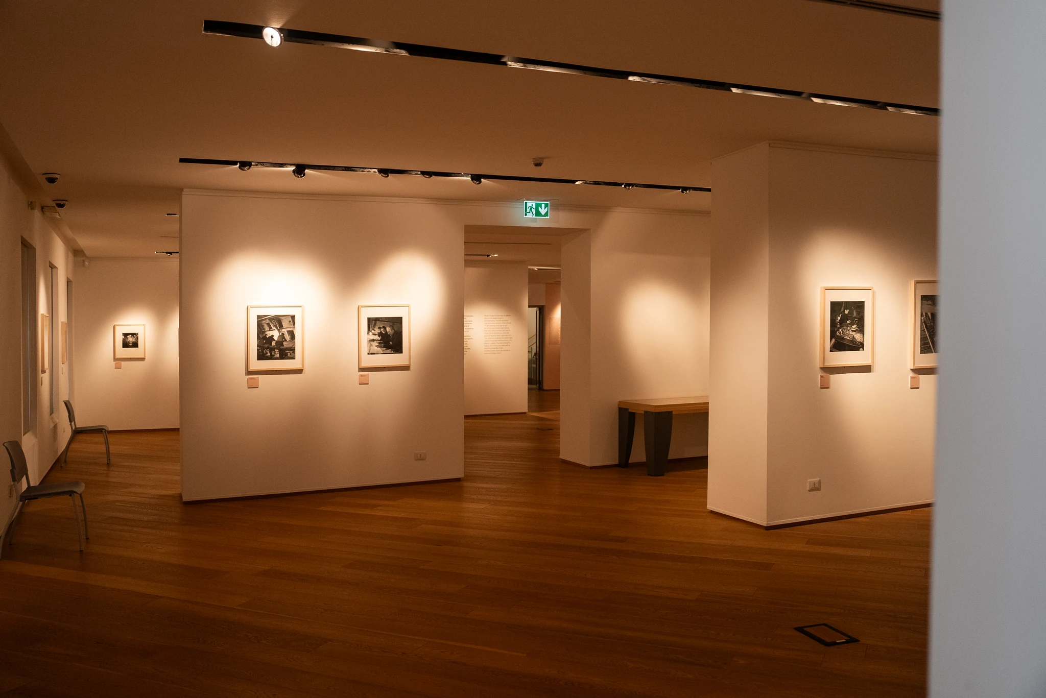 Sala espositiva con pareti bianche illuminate da faretti a soffitto. Diversi quadri incorniciati sono appesi alle pareti, ciascuno con una piccola etichetta sottostante. Il pavimento è in legno e nella stanza si aprono passaggi verso altre aree della mostra.