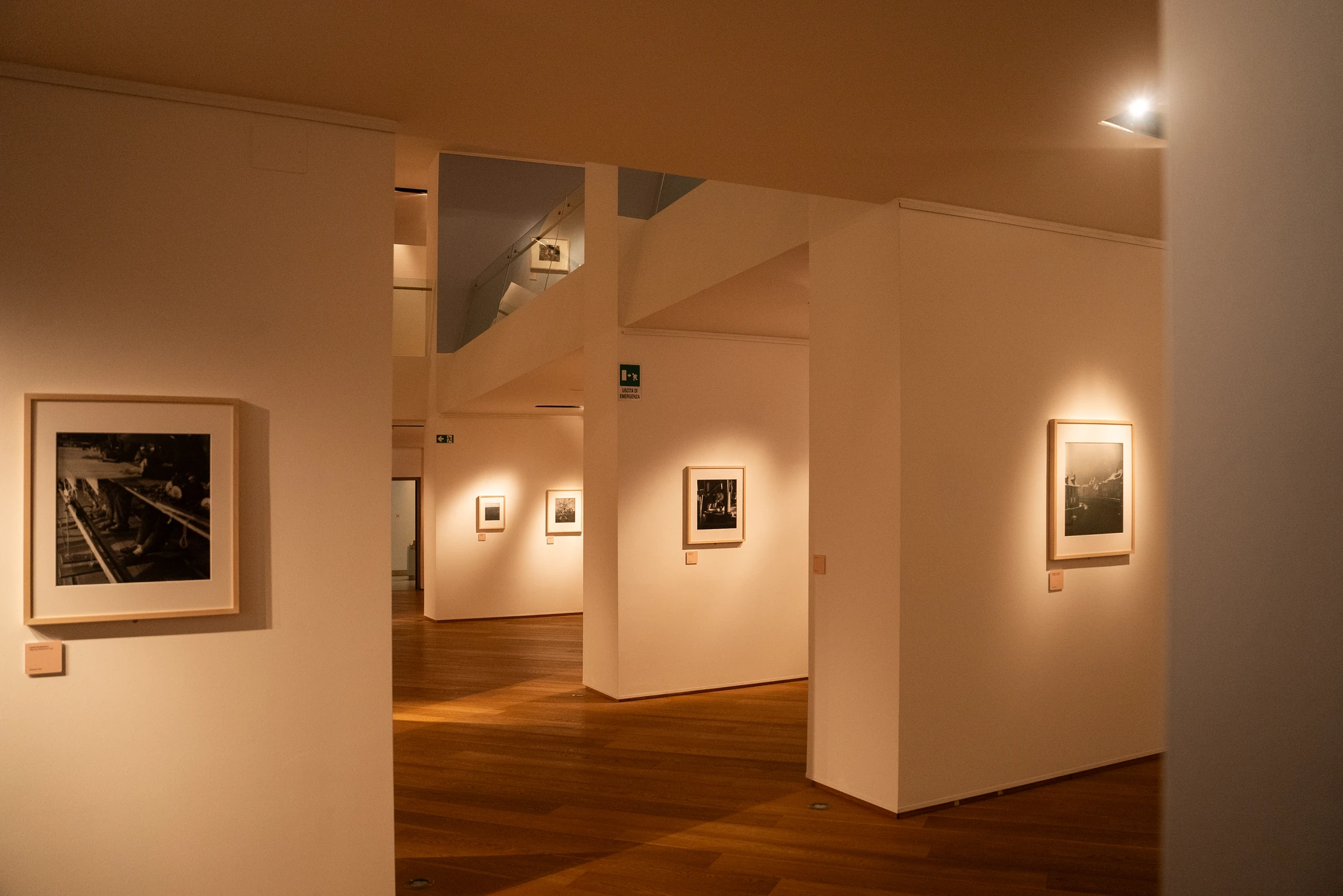 Sala espositiva con pareti bianche e pavimento in legno, illuminata da faretti a soffitto. Diversi quadri incorniciati in bianco e nero sono appesi alle pareti, ciascuno con una piccola etichetta sottostante. Le pareti creano passaggi e scorci prospettici che conducono verso altri ambienti della mostra.