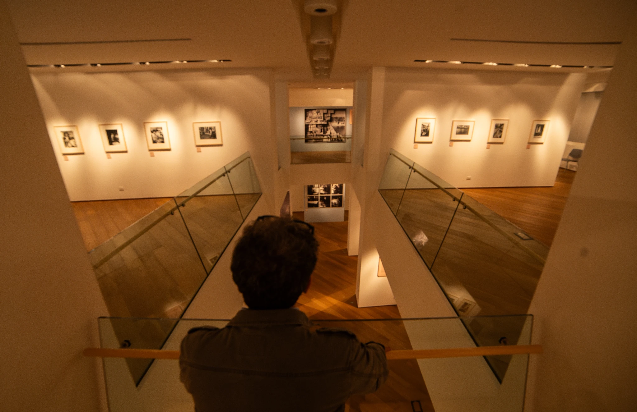 Persona vista da dietro mentre osserva una sala espositiva a due livelli, con corridoi laterali dotati di parapetti in vetro e pareti bianche decorate con fotografie incorniciate. Il pavimento è in legno e, sul fondo, è visibile un pannello con una composizione di immagini.