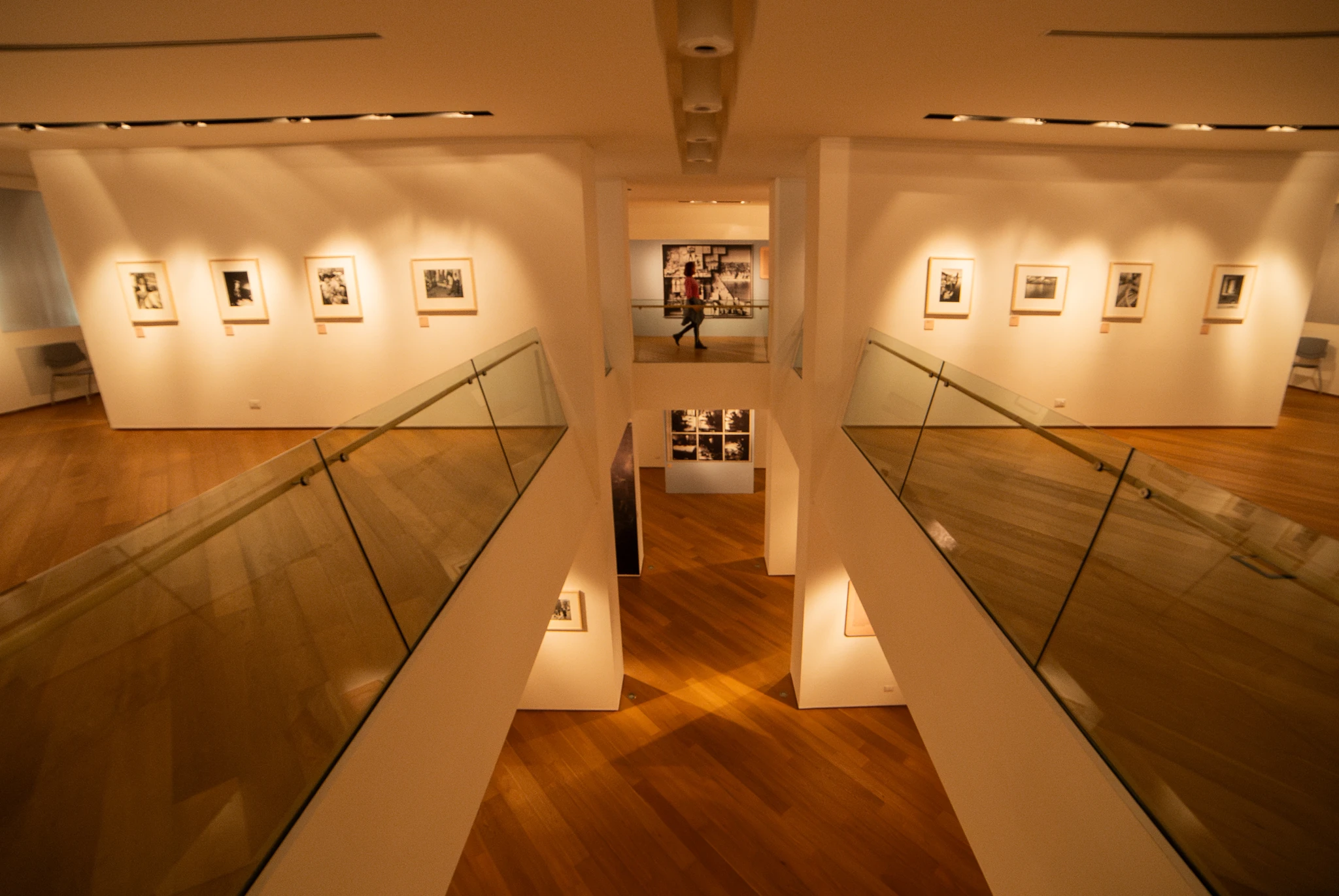 Ampio spazio espositivo a due livelli, con pavimento in legno chiaro e corridoi laterali delimitati da parapetti in vetro. Sulle pareti bianche del livello superiore sono appese fotografie incorniciate, illuminate da faretti a soffitto. In fondo, al centro, si apre una vista su un’altra sala espositiva con ulteriori immagini.