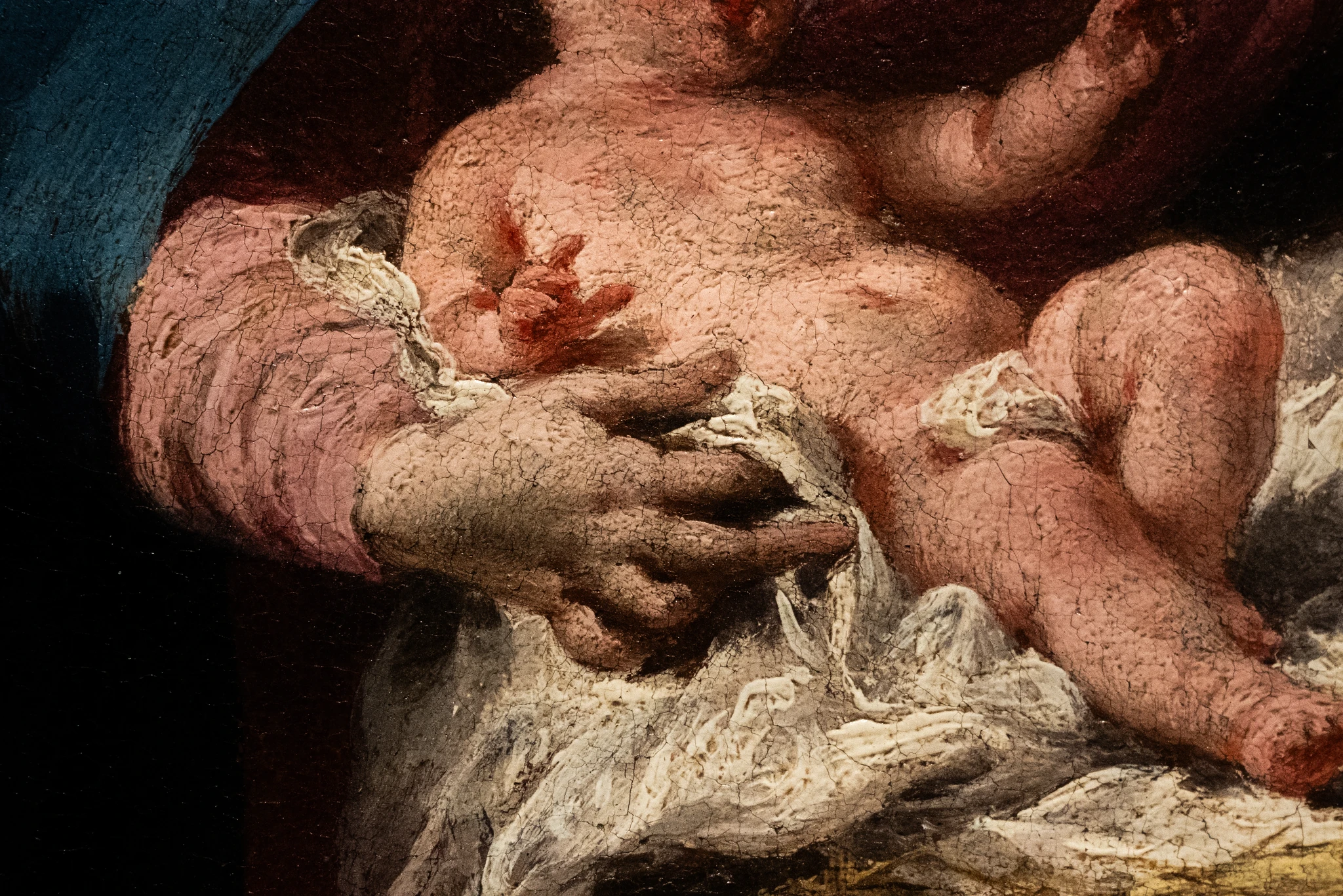 Un dettaglio di un dipinto a olio che ritrae un Gesù Bambino parzialmente nudo. La pelle del bambino è di un rosa rosato e presenta una texture craquelé visibile, che suggerisce la vetustà dell'opera. Il bambino è avvolto in un panno bianco drappeggiato e poggia su un tessuto rosso-brunastro. Una mano adulta, forse quella di Maria o di un altro personaggio, lo sorregge con delicatezza. La mano adulta è di una tonalità più scura e anche la sua pelle presenta una texture craquelé. Lo sfondo è scuro e indistinto, concentrando l'attenzione sul bambino e sulla mano. Lo stile è realistico e ricco di dettagli materici.