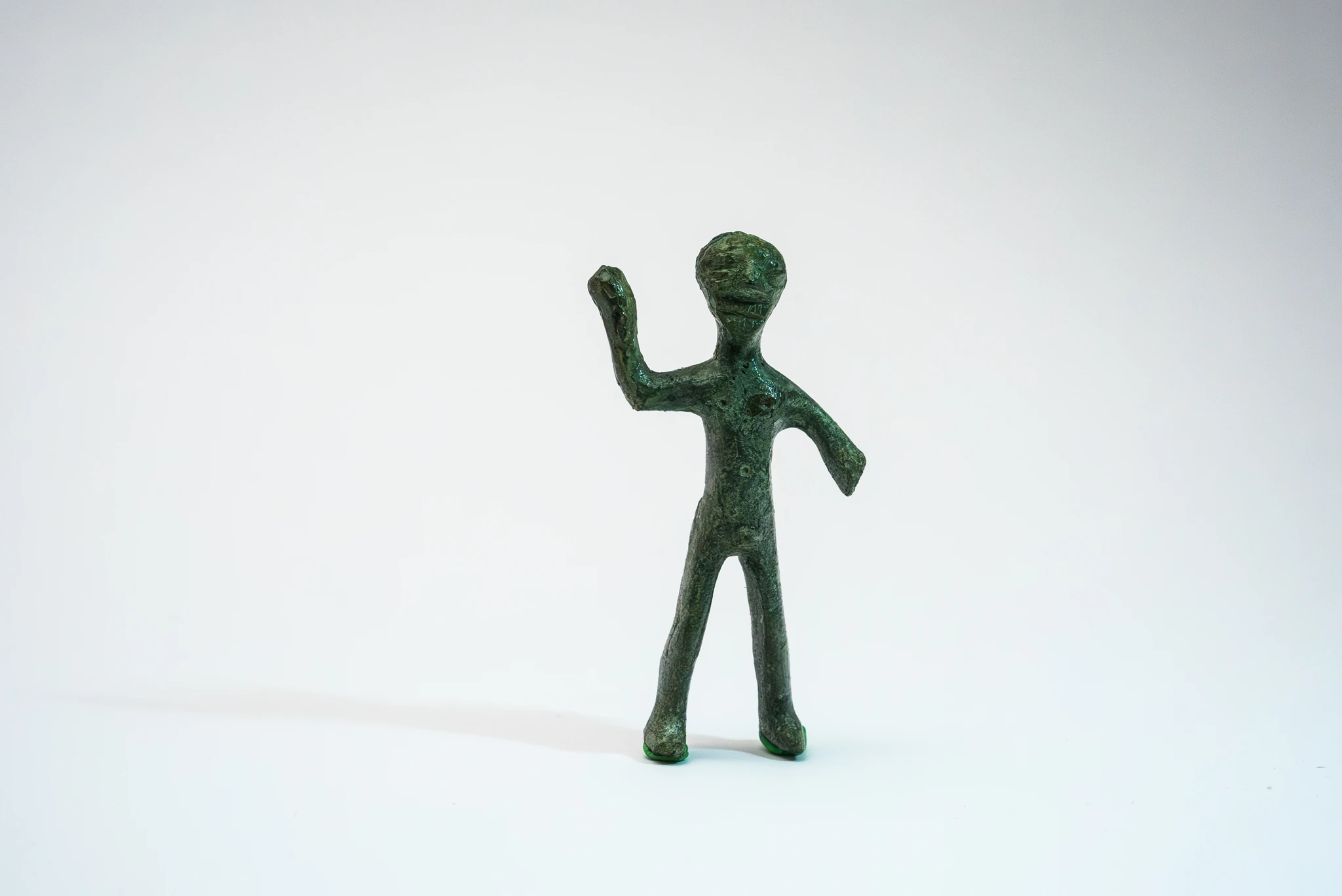 Piccola statuetta antropomorfa in bronzo stilizzata, con corpo allungato e arti semplificati. La figura è in posizione eretta, con un braccio sollevato e l’altro inclinato verso il basso. Lo sfondo è neutro e chiaro.