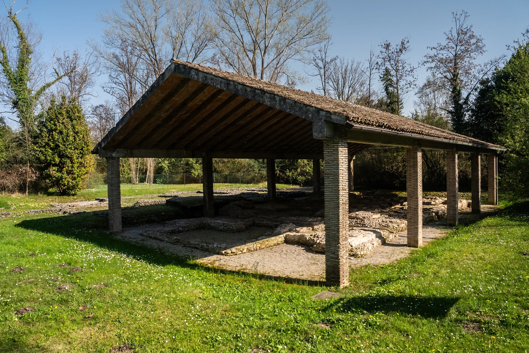 Area archeologica all’aperto con un ampio tetto a falde sostenuto da pilastri in mattoni. Sotto la struttura si trovano resti di mura e pavimentazioni antiche in pietra. L’area è circondata da prato e alberi ad alto fusto, illuminati dalla luce del giorno