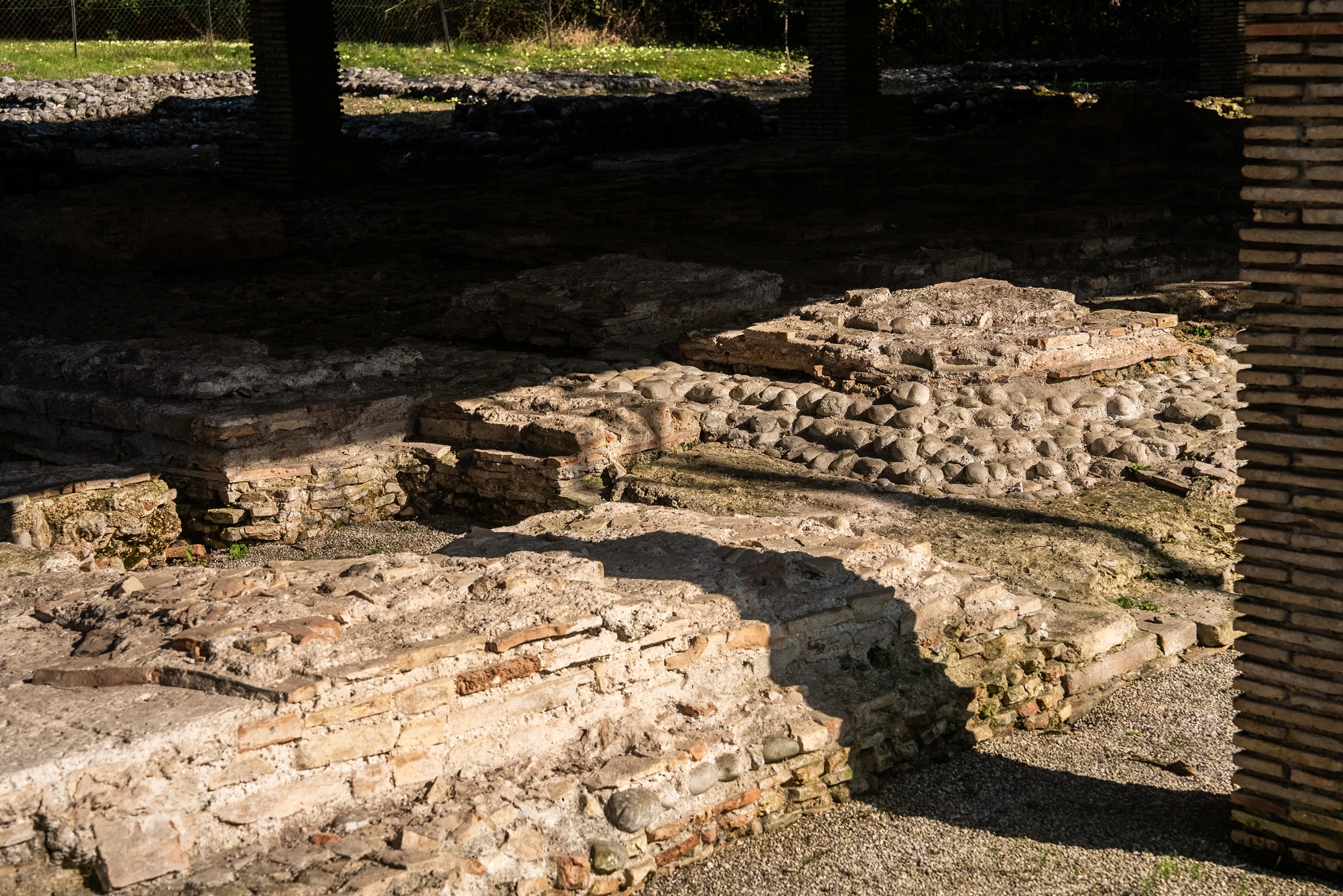 Area archeologica all’aperto con resti di strutture murarie in pietra e mattoni. In primo piano si distinguono basamenti e superfici irregolari composte da ciottoli e frammenti lapidei. La luce solare crea zone illuminate e ombreggiate tra i resti, mentre sullo sfondo si intravedono vegetazione e parti di un recinto.