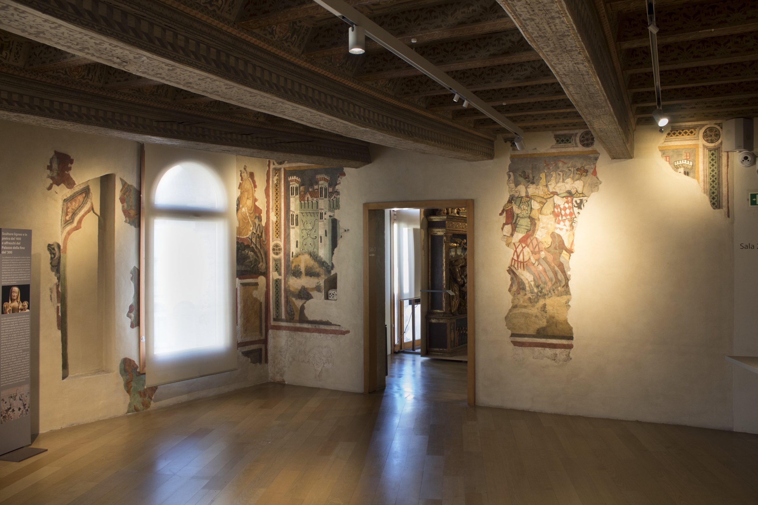Interno di una sala museale con pavimenti in legno e soffitto a cassettoni decorato. Sulle pareti chiare sono visibili frammenti di affreschi medievali che ritraggono un castello e scene di cavalieri. Una finestra ad arco sulla sinistra è coperta da una tenda bianca che diffonde la luce naturale.