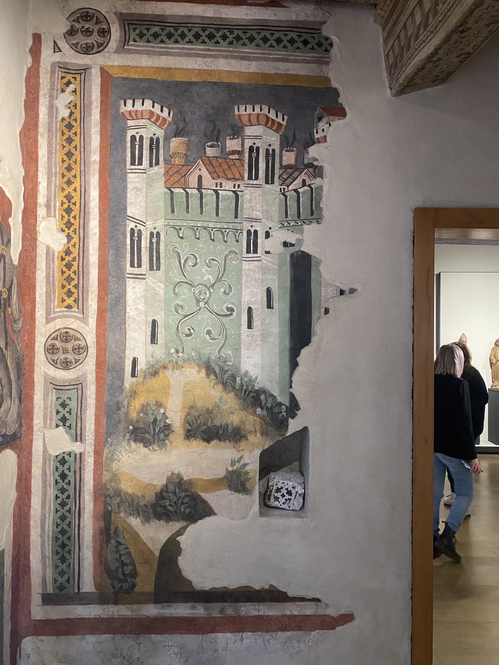 Frammento di affresco medievale su una parete museale che raffigura un castello stilizzato con alte torri bianche, merlature e tetti rossi. La facciata del castello è decorata con motivi a viticci bianchi su fondo verde. Sulla sinistra è presente una cornice decorativa verticale con motivi geometrici e floreali. A destra, una porta aperta lascia intravedere dei visitatori in una sala adiacente.
