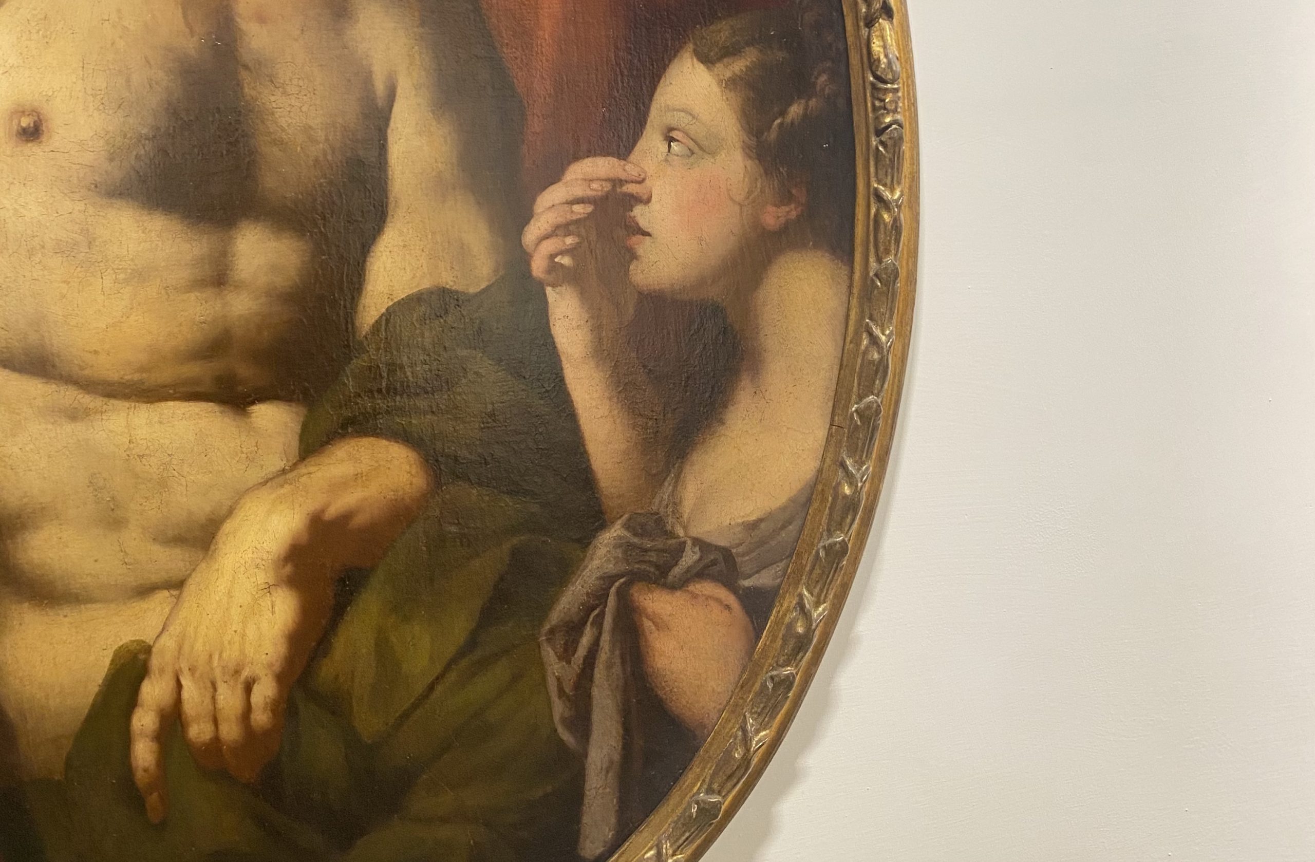 Dettaglio di un dipinto a olio di forma ovale racchiuso in una cornice dorata intagliata. L'opera raffigura una giovane donna di profilo che si porta una mano al naso con un'espressione curiosa o sorpresa. Accanto a lei, si vede il torso muscoloso di una figura maschile parzialmente coperta da un drappo verde scuro. Lo stile pittorico è classico, con un uso morbido del chiaroscuro tipico del XVII o XVIII secolo.
