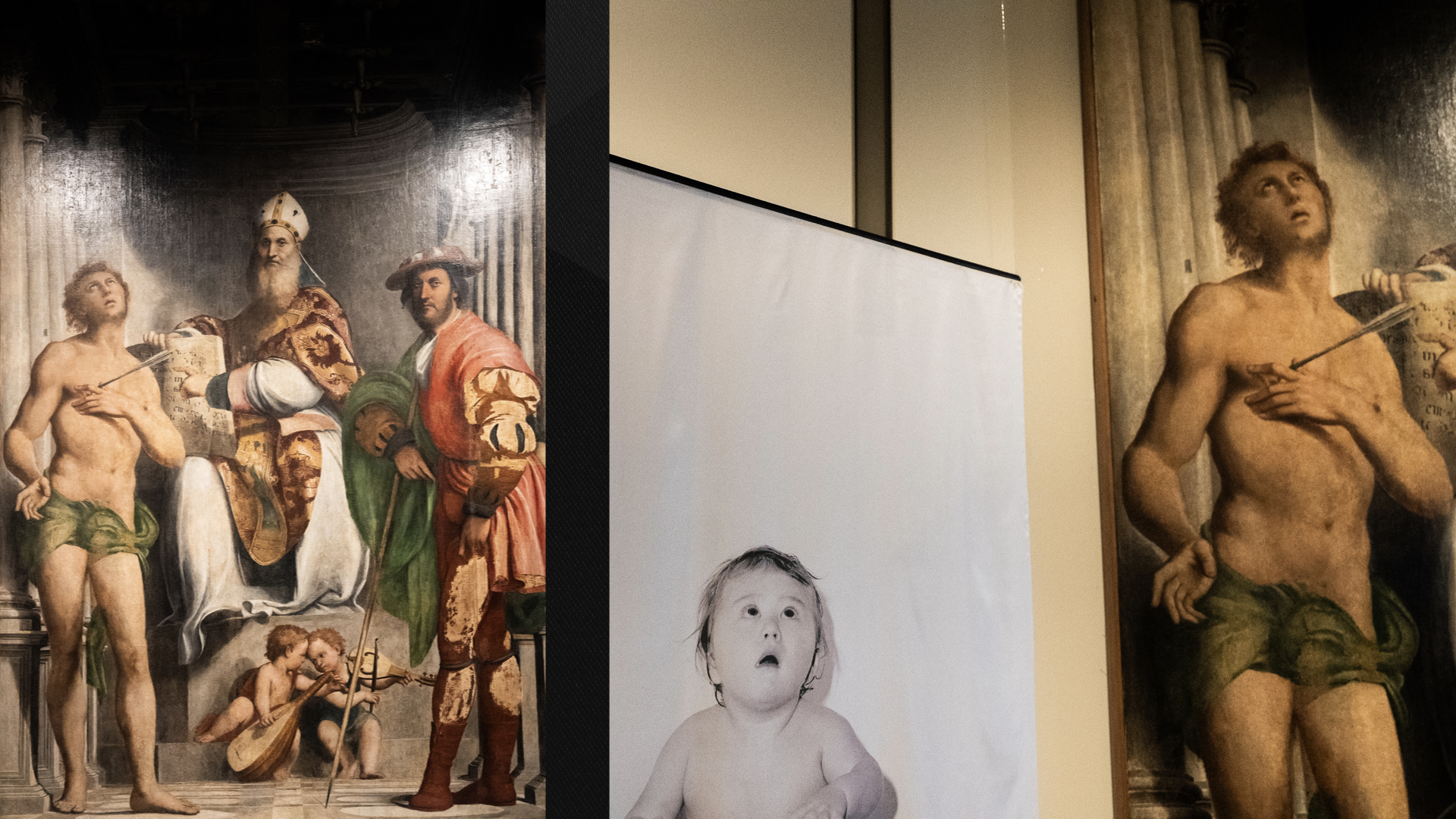 Due immagini affiancate che creano un parallelismo visivo. A sinistra: Un dipinto rinascimentale raffigurante una "Sacra Conversazione". Al centro un vescovo in trono, a sinistra San Sebastiano trafitto da una freccia e a destra un nobile o un santo in vesti rinascimentali rosse. Ai piedi del trono, due piccoli angeli musicanti. A destra: Un dettaglio ravvicinato che accosta il San Sebastiano del dipinto alla fotografia in bianco e nero di un bambino che guarda verso l'alto con un'espressione di stupore, richiamando la posa e lo sguardo rivolto al cielo del santo.