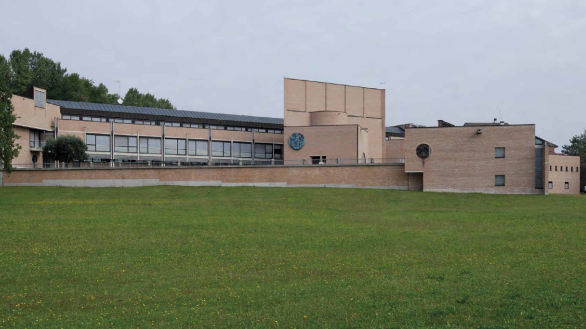 Veduta esterna panoramica del Centro Diocesano di Pordenone, sede del Museo Diocesano. L'edificio moderno in mattoni a vista presenta geometrie rigorose e un'ampia facciata con vetrate continue, situato davanti a un vasto prato verde sotto un cielo nuvoloso.