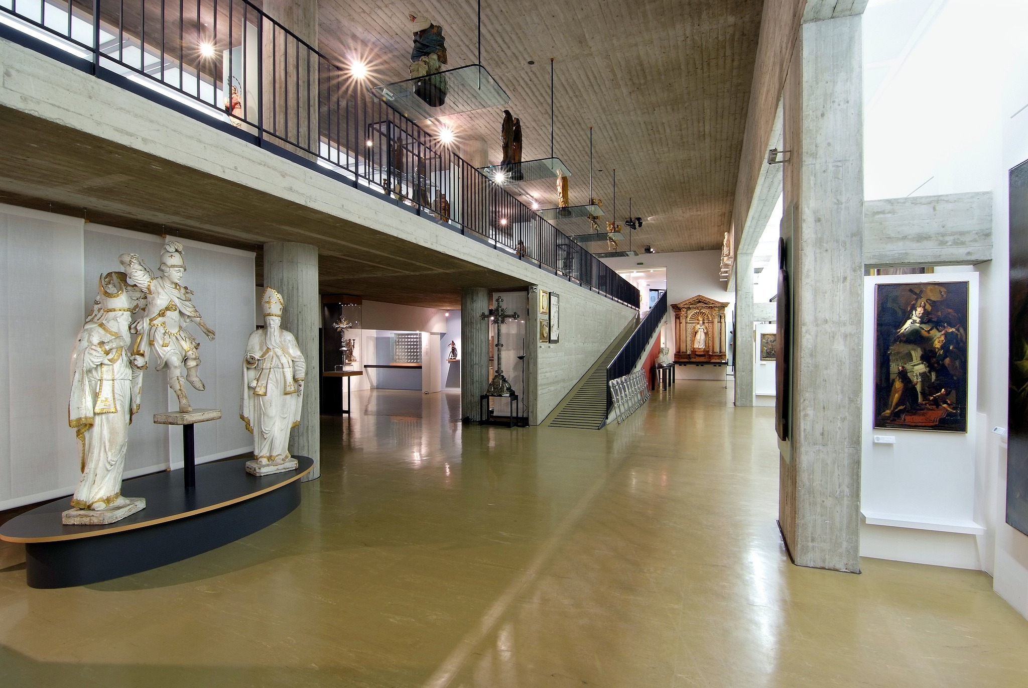 Una fotografia dell'interno di un museo d'arte sacra dal design moderno e industriale. Lo spazio è ampio, con pavimenti lucidi in resina beige e strutture in cemento armato a vista. Sulla sinistra, in primo piano, tre grandi statue lignee dipinte di bianco e oro sono esposte su una base scura. Al centro della sala, una rampa e una scala conducono a un ballatoio superiore protetto da una ringhiera metallica nera, dove altre sculture sono esposte su mensole di vetro sospese. Sulla destra, un pilastro in cemento ospita un dipinto incorniciato. In fondo alla sala si scorgono ulteriori opere d'arte, tra cui un altare ligneo dorato e una croce processionale. L'illuminazione è puntuale, con faretti che mettono in risalto i singoli pezzi esposti.